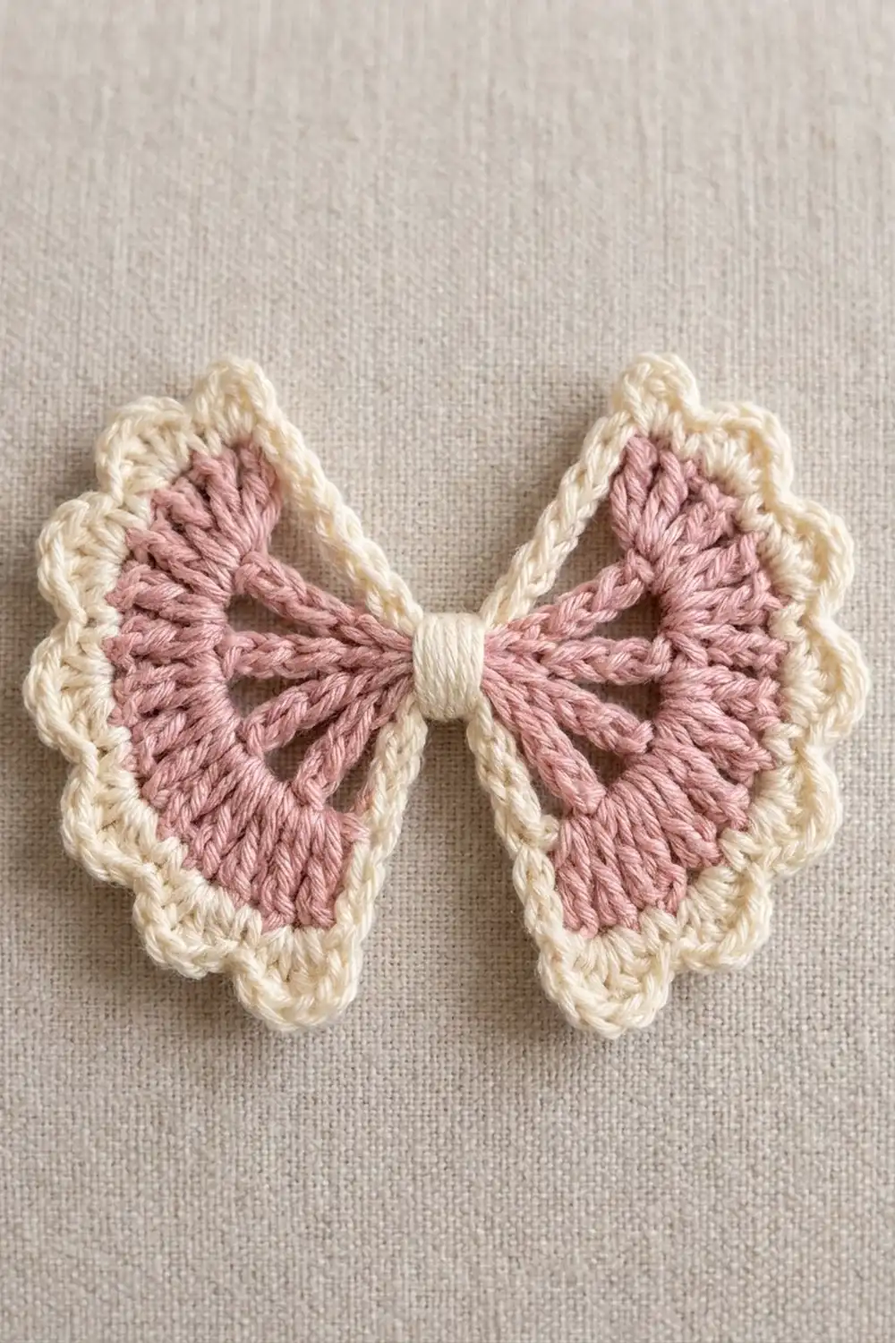 A delicate crocheted butterfly with pink wings and cream-colored borders on a beige linen fabric background.