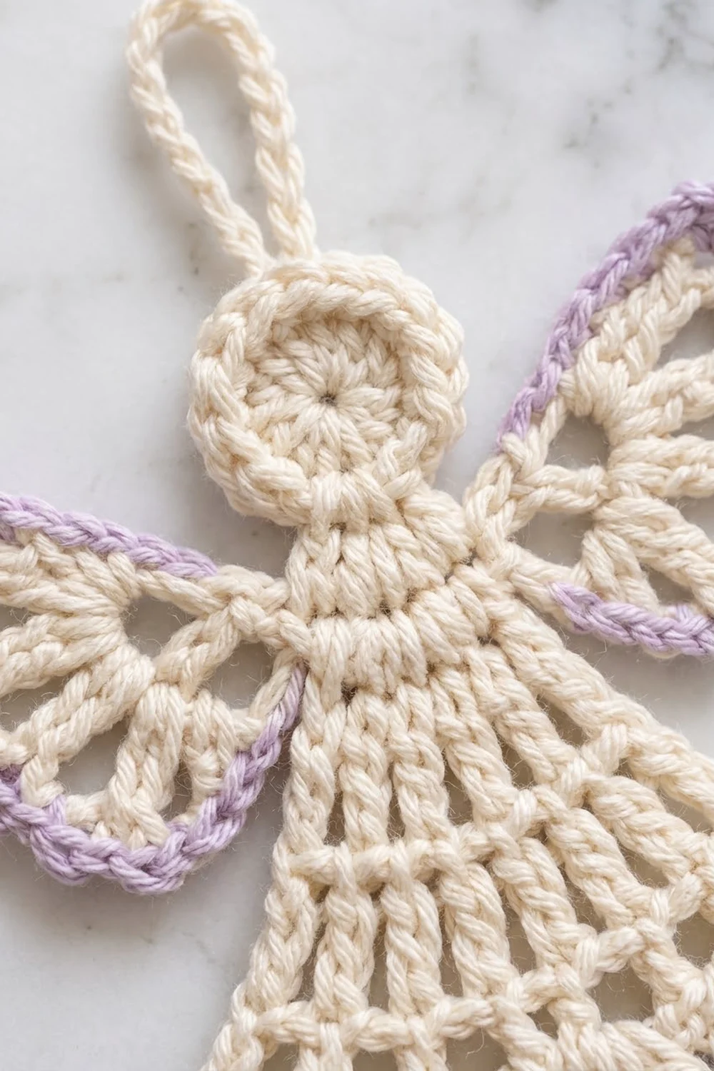 A handmade macramé ornament featuring a cream-colored knotted figure with lavender-accented arms, displayed on marble.