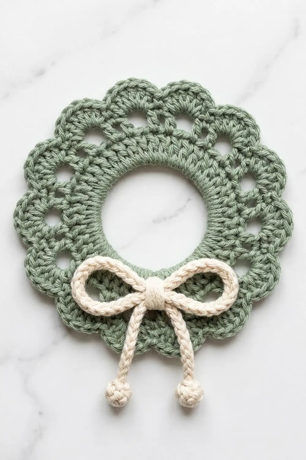 A crocheted wreath in sage green with a cream-colored bow and knotted ties on white marble.