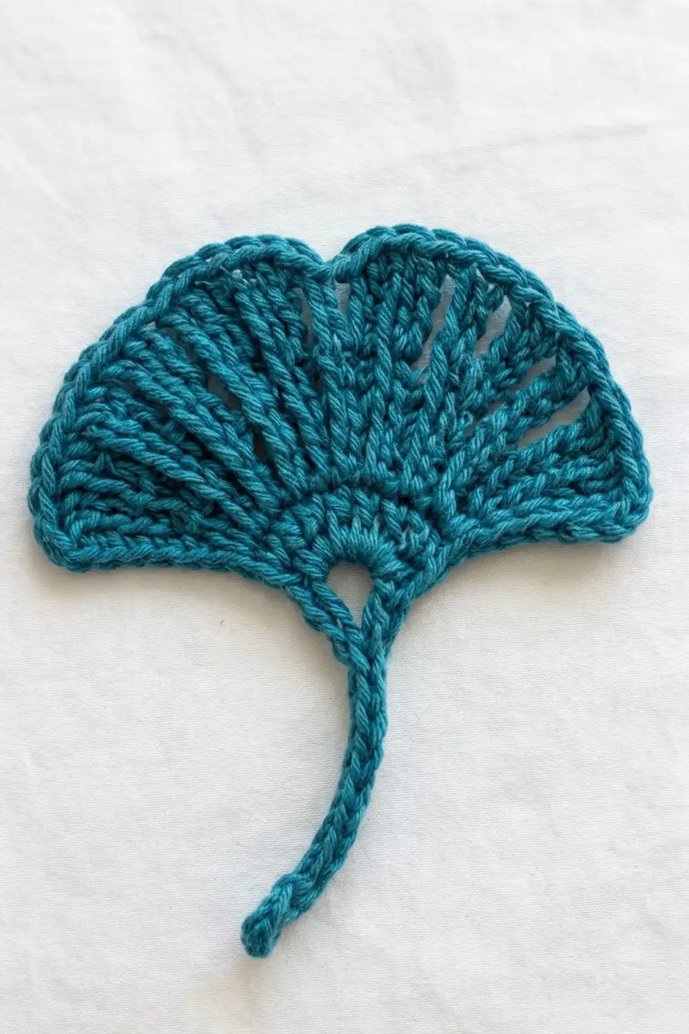 Teal crocheted ginkgo leaf with a twisted stem displayed flat against a white fabric background.