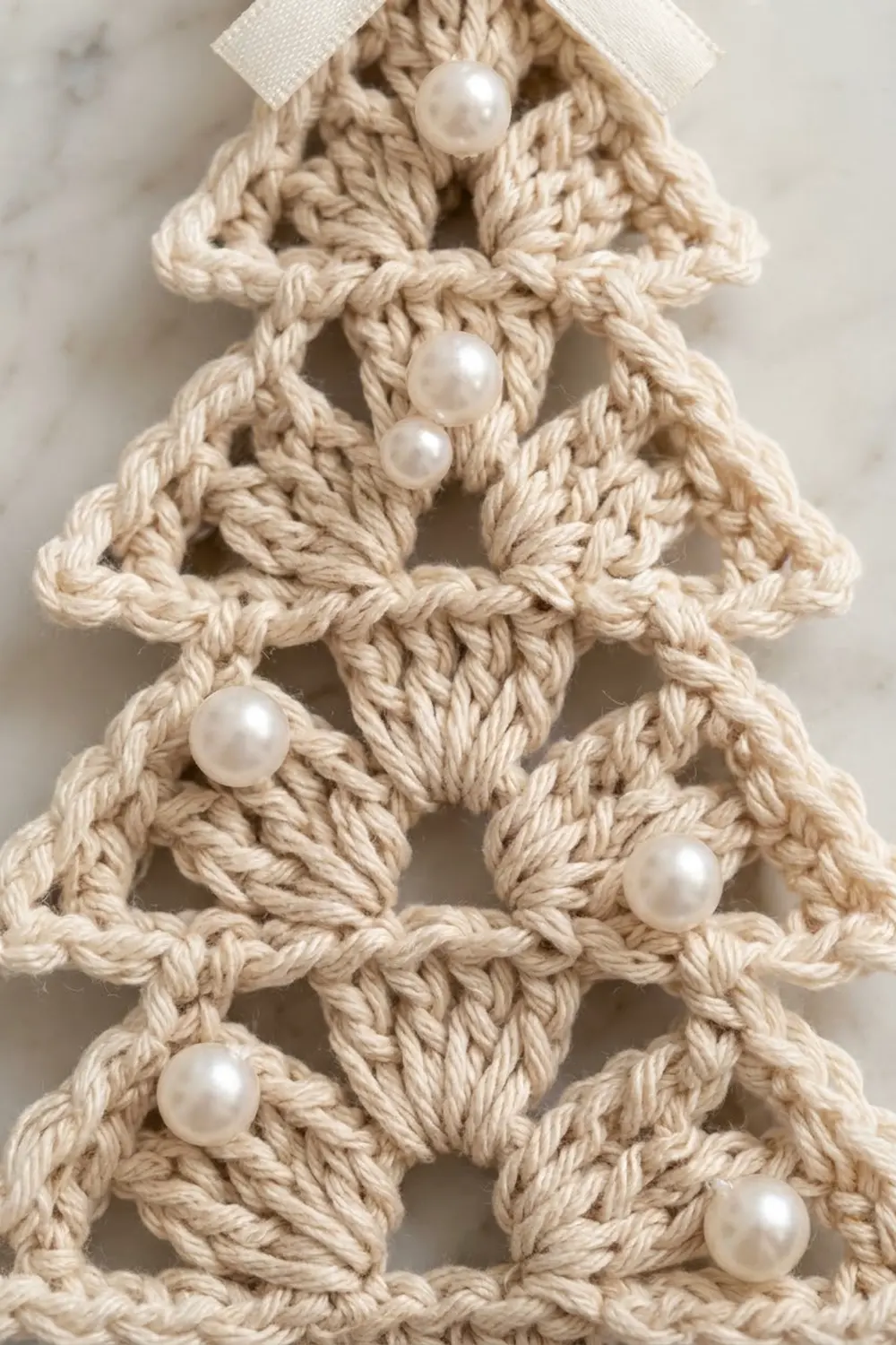A macramé Christmas tree ornament in cream rope with pearl beads and a white ribbon bow against marble.