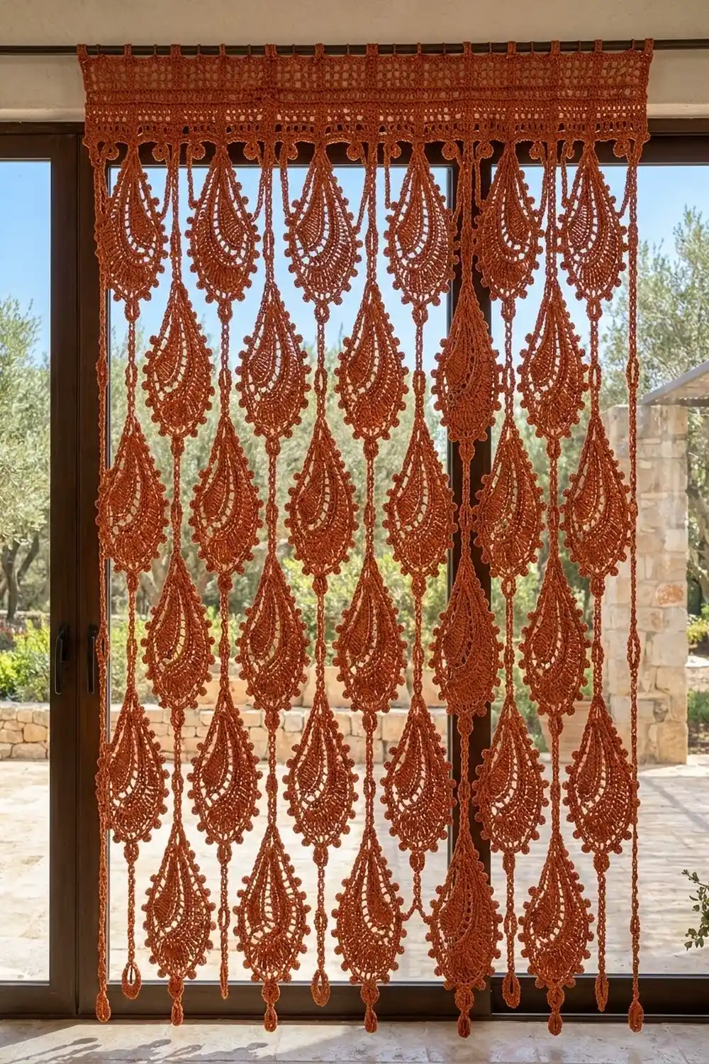 Rust-colored macramé curtain with intricate teardrop patterns hanging over a glass door overlooking a garden.