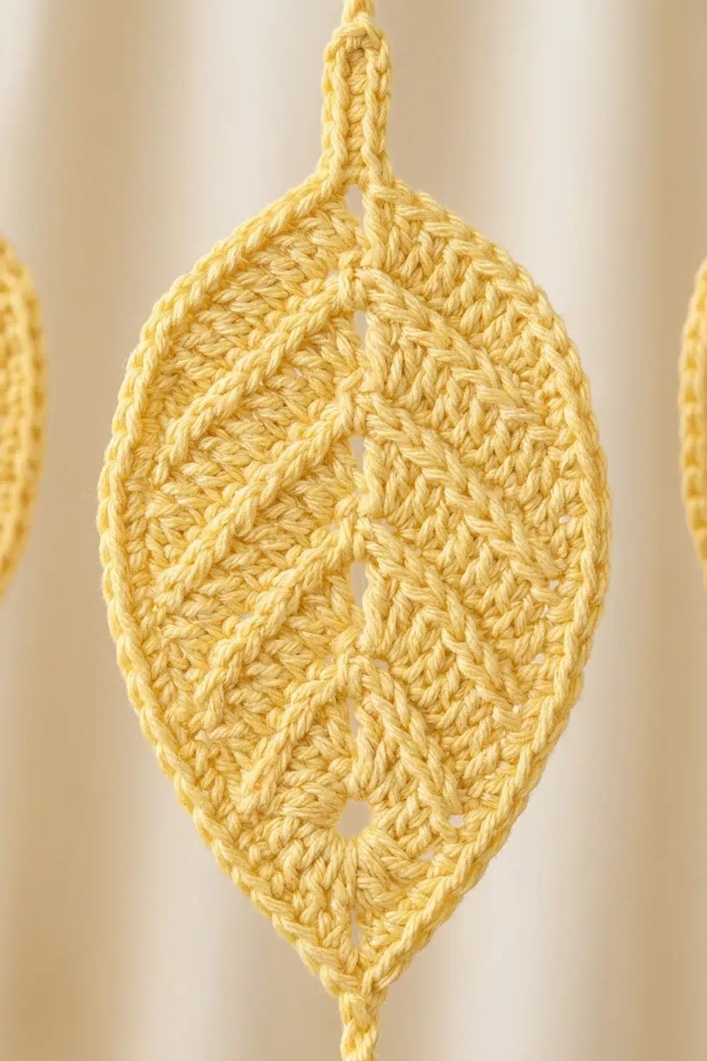 A crocheted golden-yellow leaf ornament with an open lace pattern hangs against a soft, blurred cream background.