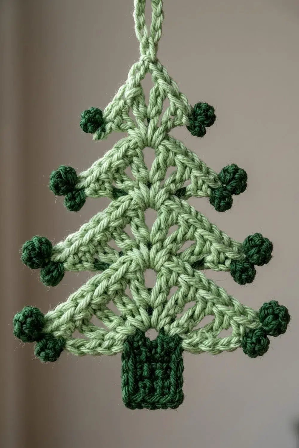 Handmade crocheted Christmas tree ornament in light and dark green yarn with pom-pom decorations hanging against a neutral background.