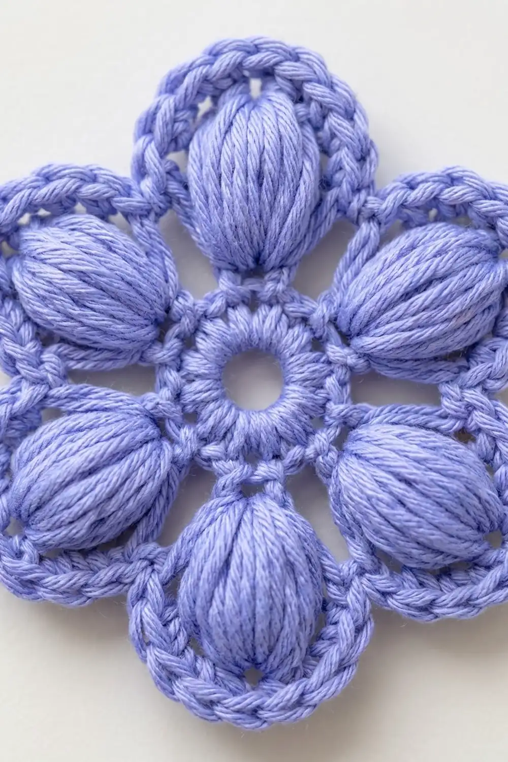 Periwinkle blue crocheted flower with braided petals and openwork center detail against white background.