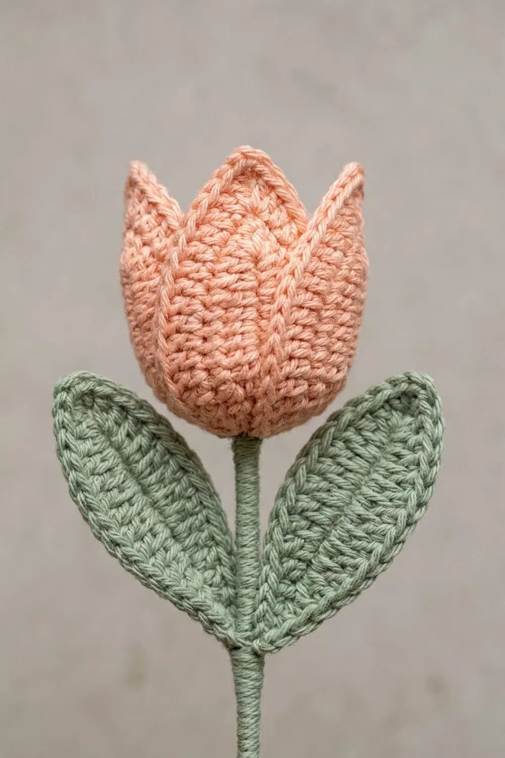 A crocheted tulip with a pink bloom and green leaves against a neutral gray background.
