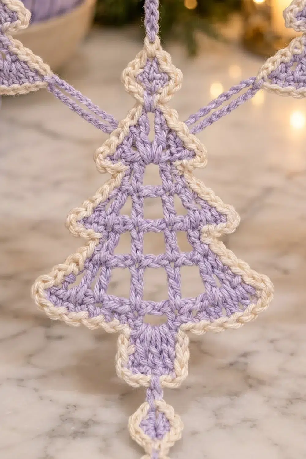 A crocheted purple and cream Christmas tree ornament with openwork pattern hangs against a blurred festive background.