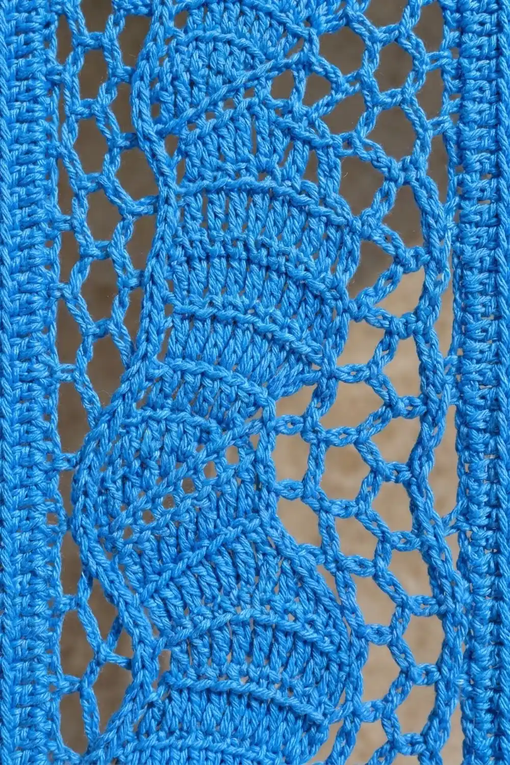 Close-up of bright blue crocheted lace fabric with open mesh pattern and diamond-shaped holes against neutral background.