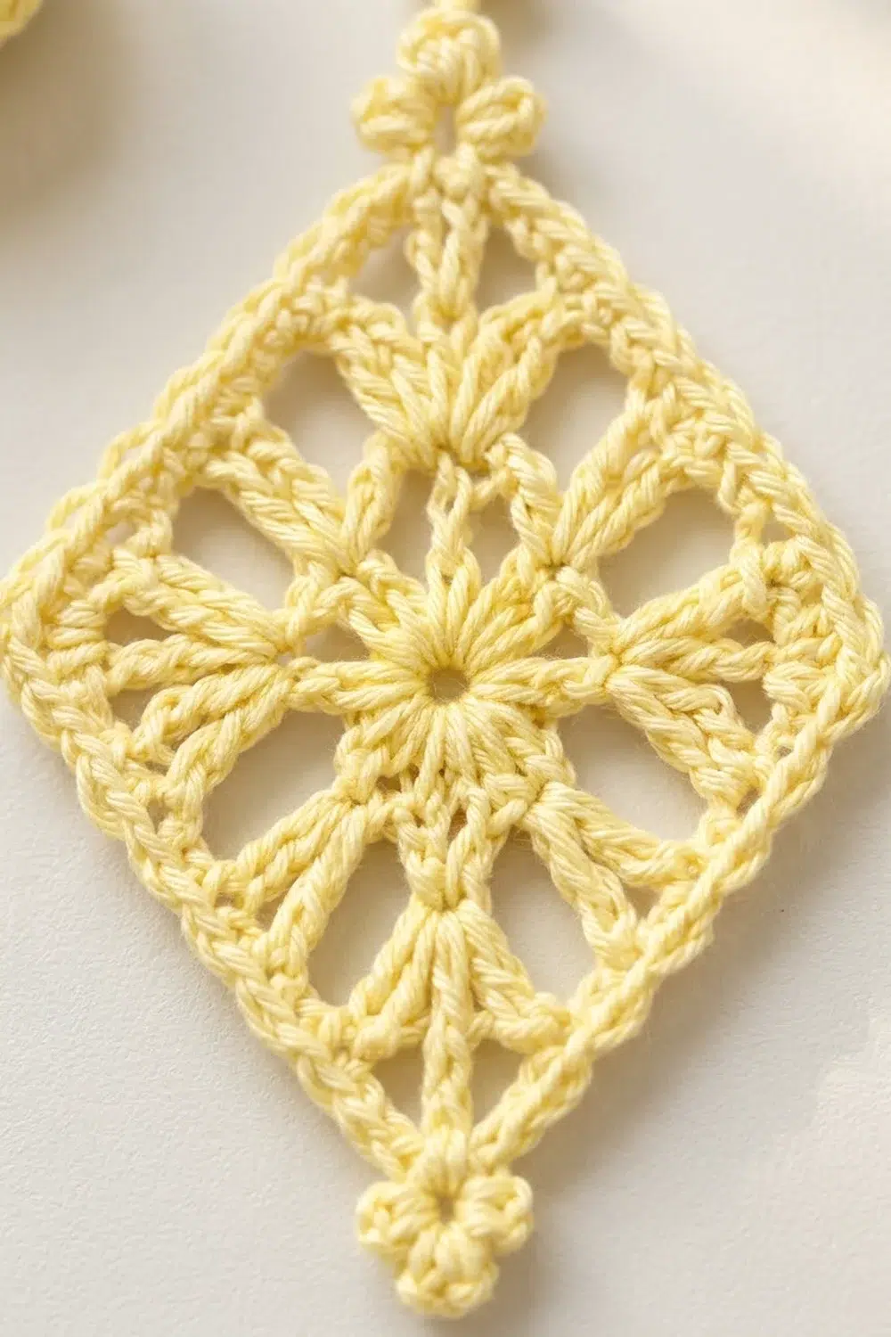 A pale yellow crocheted diamond-shaped ornament with intricate lacework pattern displayed against a white background.