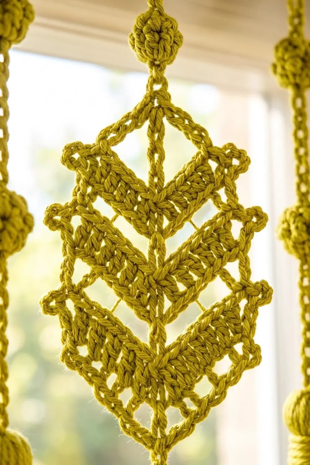 A mustard yellow crocheted diamond ornament hangs from a chain against a blurred indoor background.