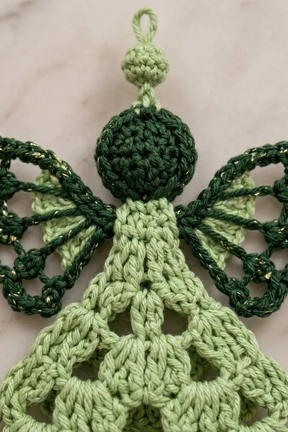 Crocheted green and light green angel ornament with braided rope arms displayed on marble surface.