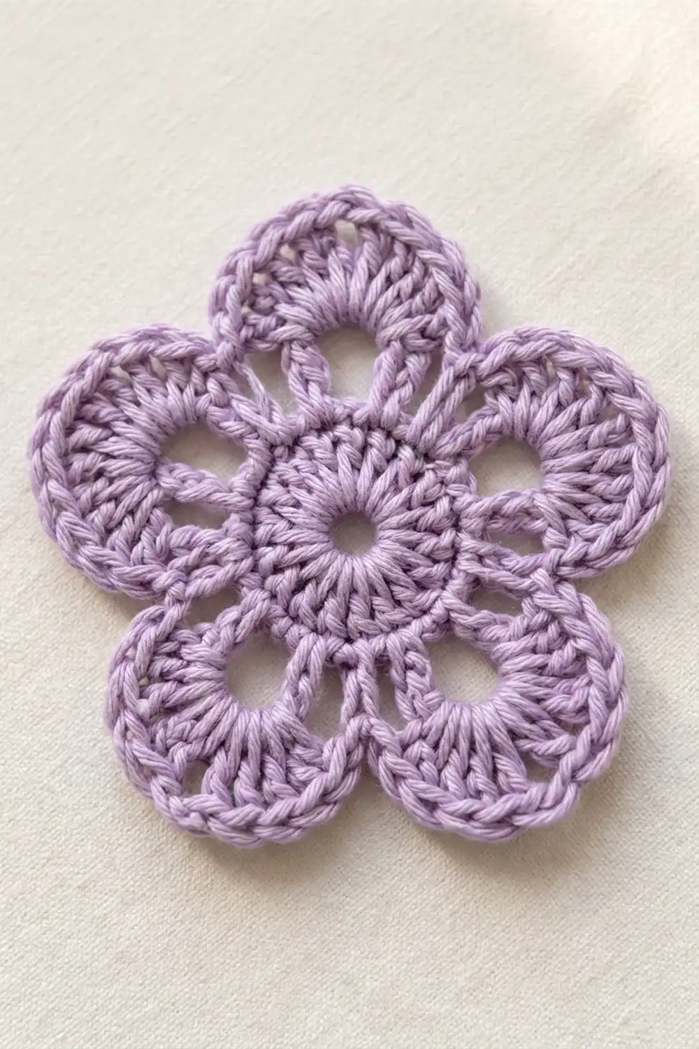 A delicate lavender crochet flower with five petals arranged symmetrically on a cream-colored textured background.