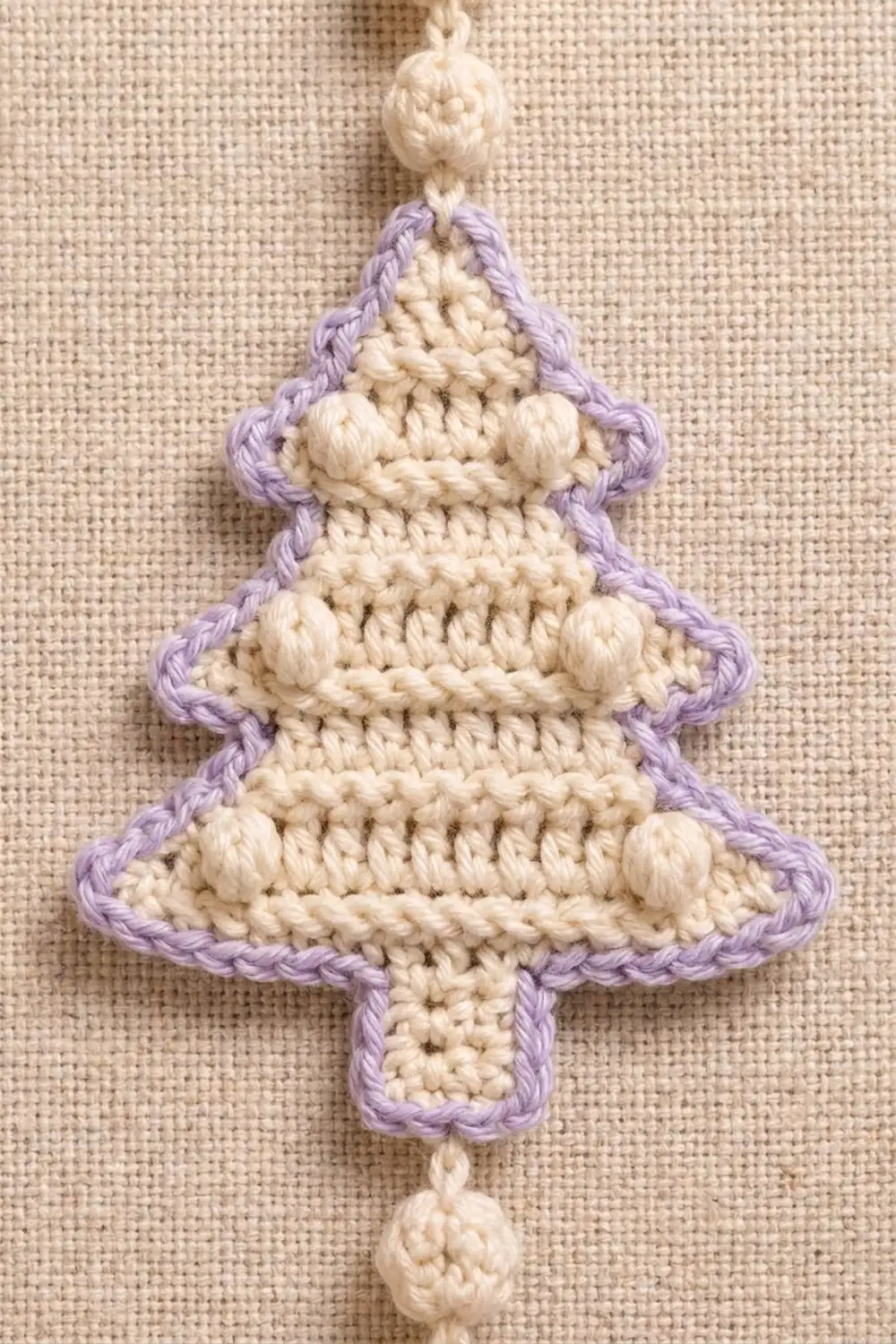 Crocheted Christmas tree ornament with lavender edging, cream interior, and beaded details on textured fabric background.
