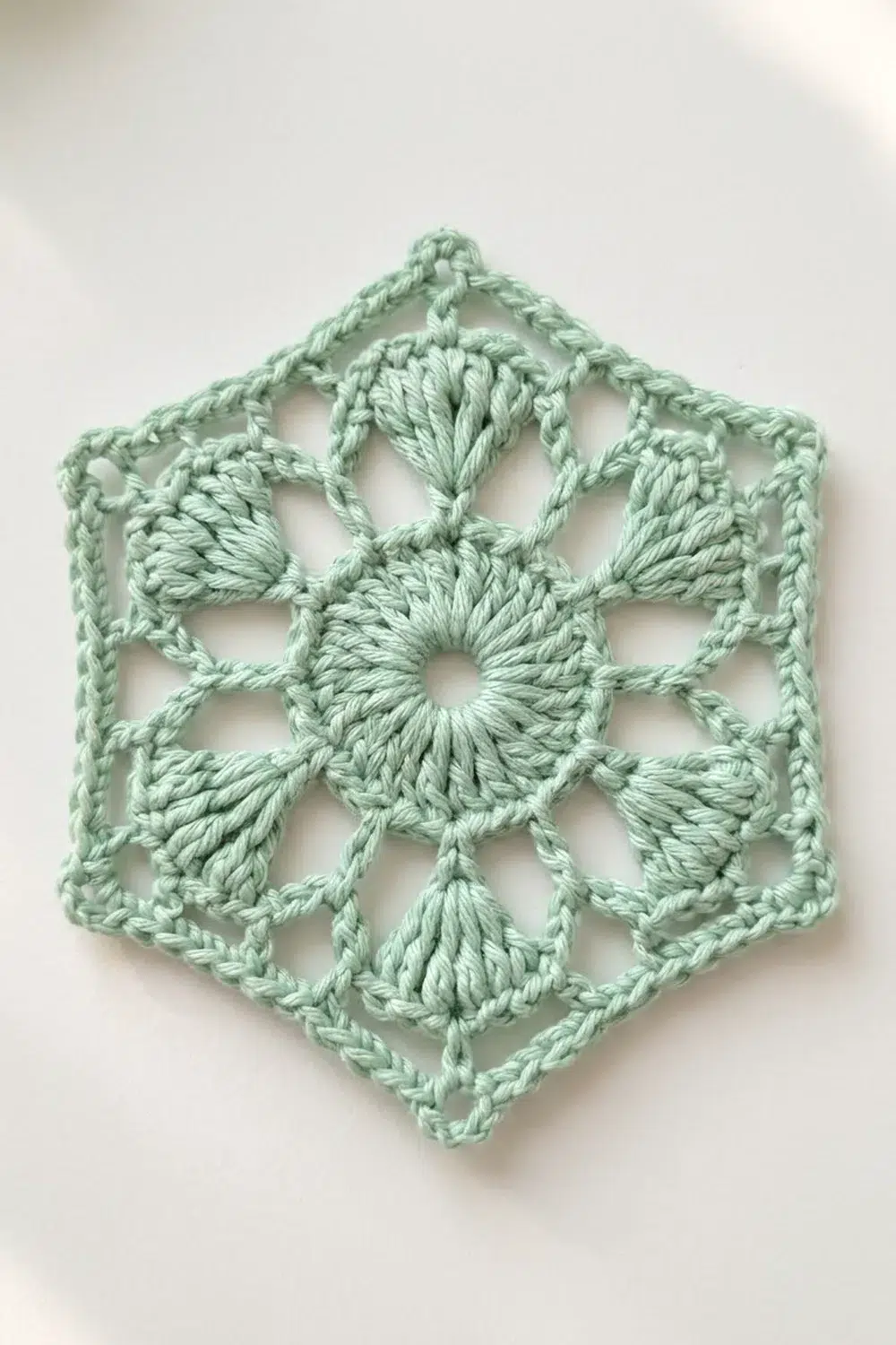 A pale green crocheted hexagonal snowflake ornament with intricate shell stitch details displayed against a white background.