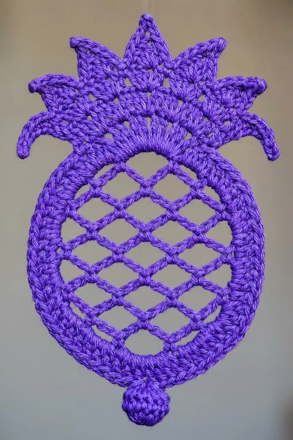 A purple crocheted pineapple appliqué with an open lattice body and decorative crown on a neutral background.