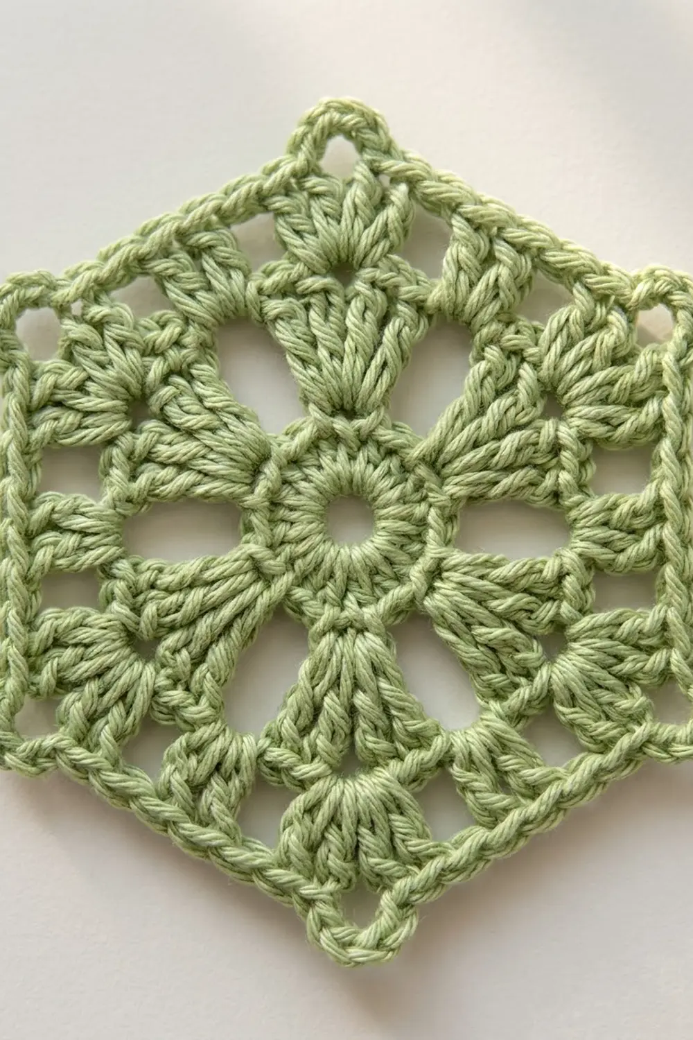 Close-up of a light green crocheted diamond-shaped ornament with intricate openwork pattern against a white background.