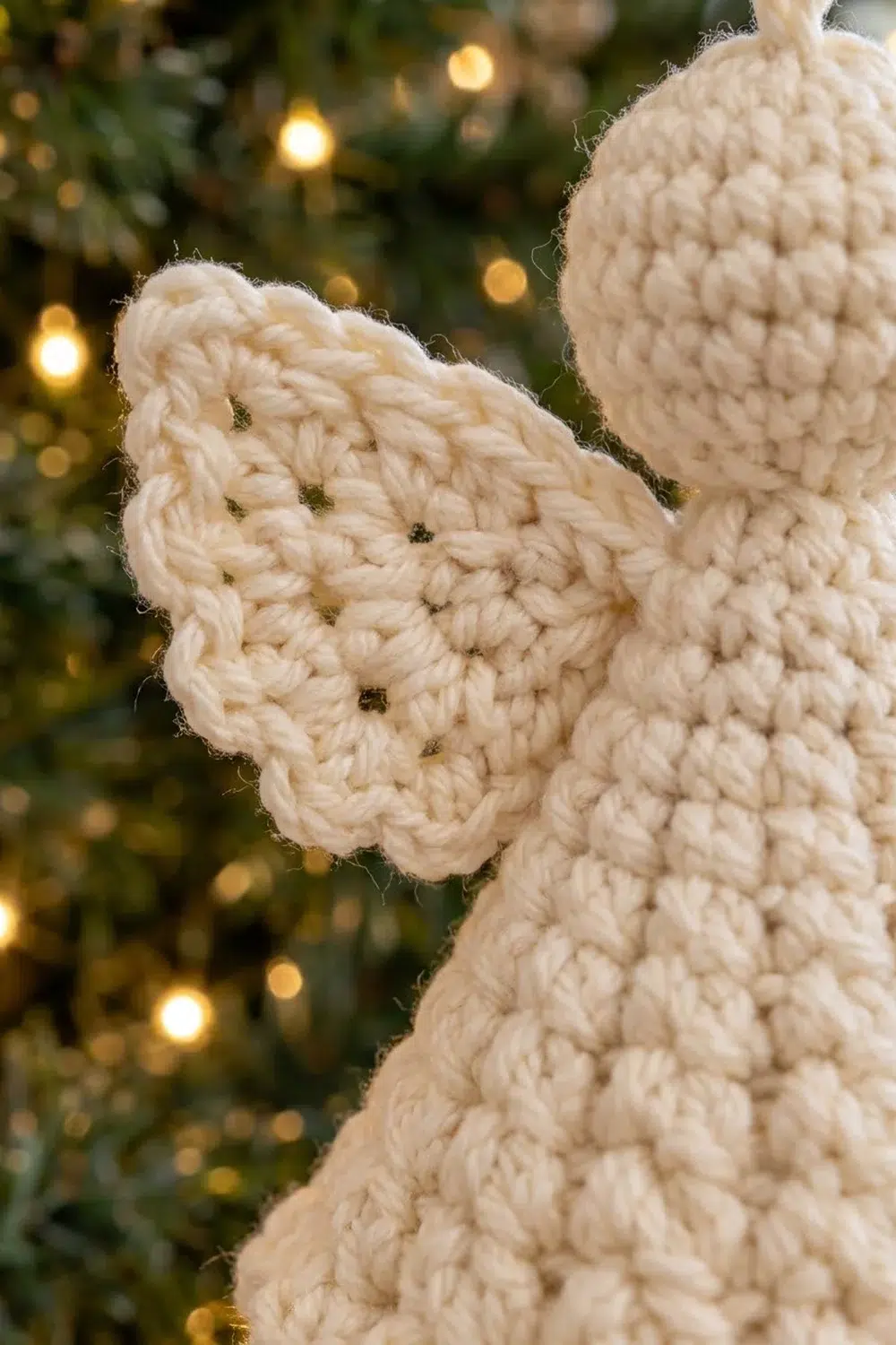 Handmade crocheted cream-colored angel ornament with wings displayed against blurred holiday lights and greenery.