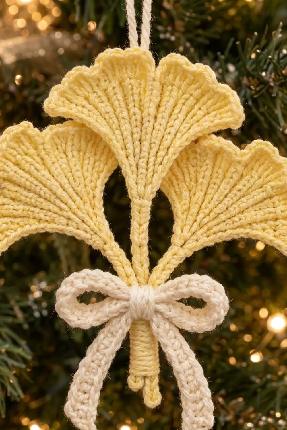 A knitted yellow flower ornament with a cream-colored bow hanging on an evergreen Christmas tree.
