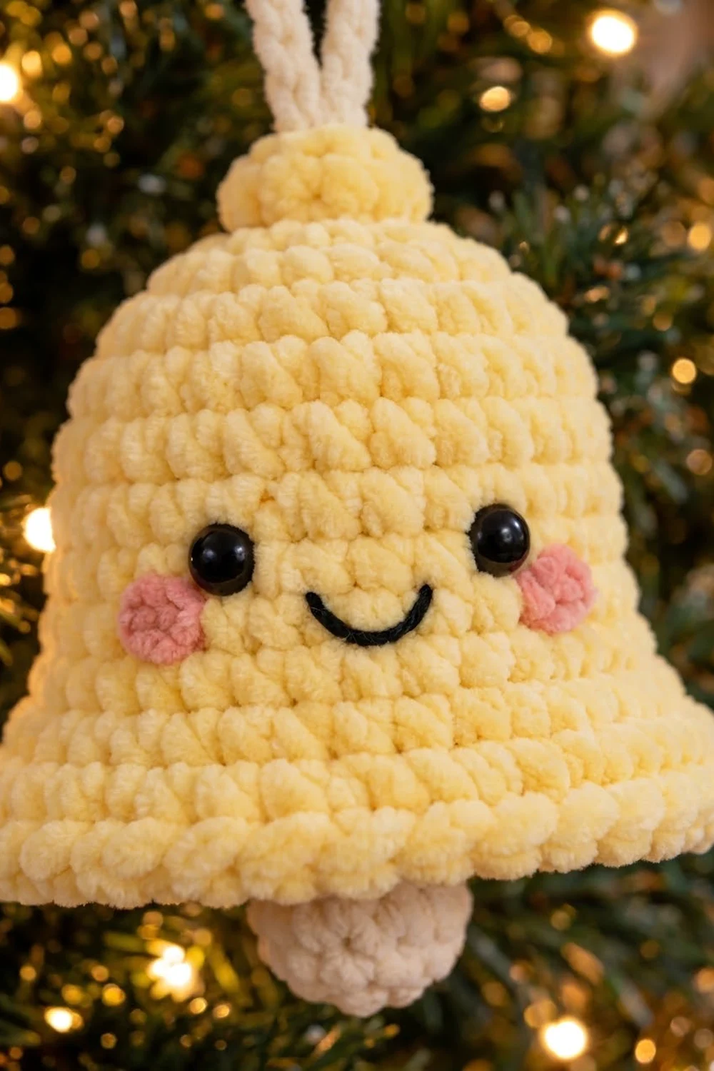 A crocheted yellow bell ornament with a smiling face hangs from a white string on a decorated Christmas tree.