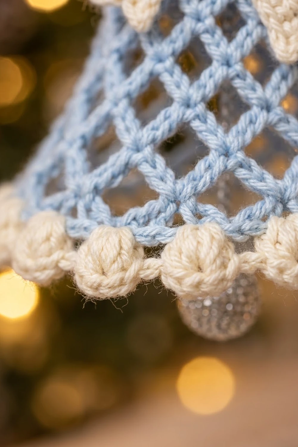 Close-up of crocheted lace with light blue and cream yarn showing detailed knot patterns against a blurred warm-lit background.