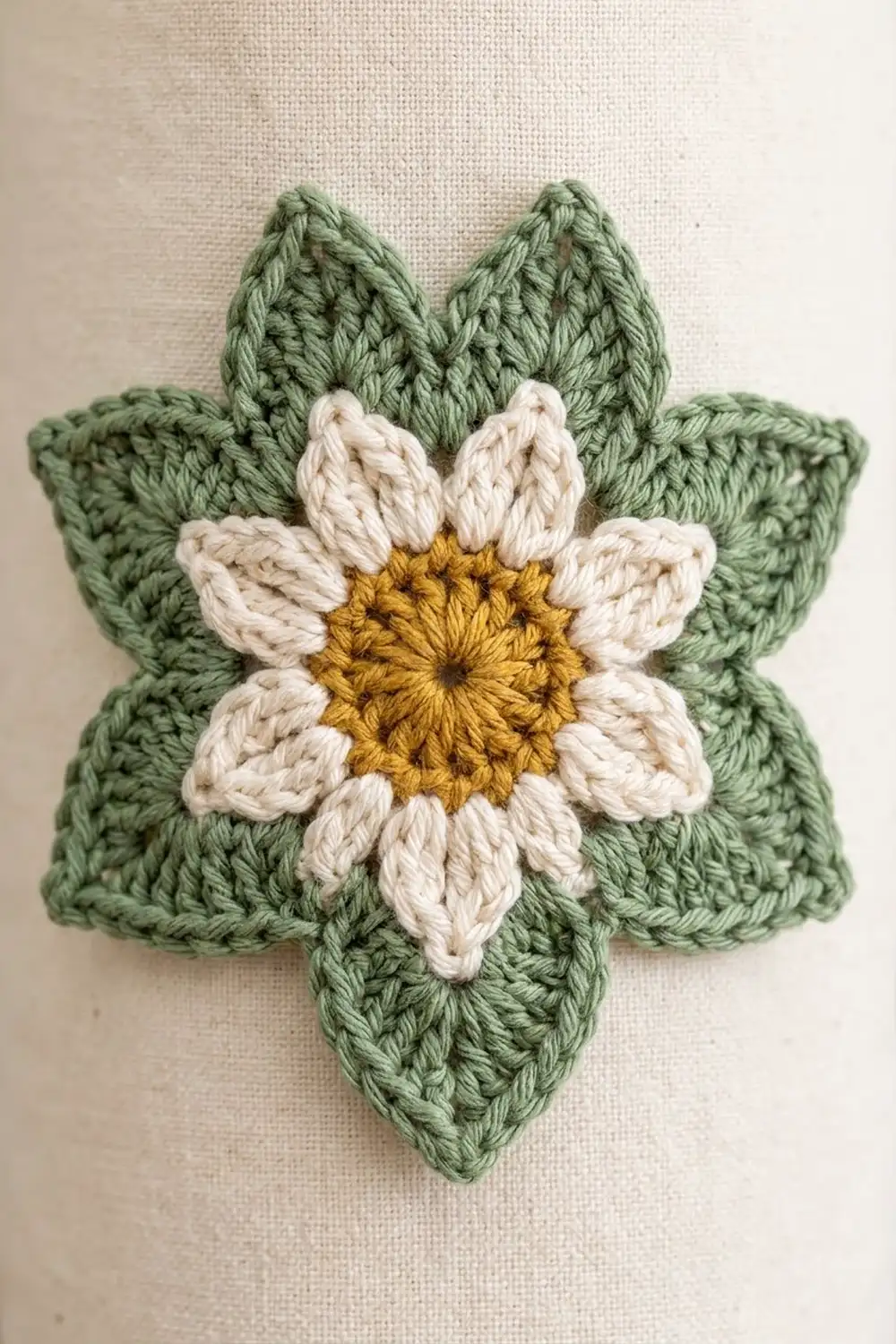 Crocheted daisy flower with cream petals, golden yellow center, and green leaves on neutral fabric background.