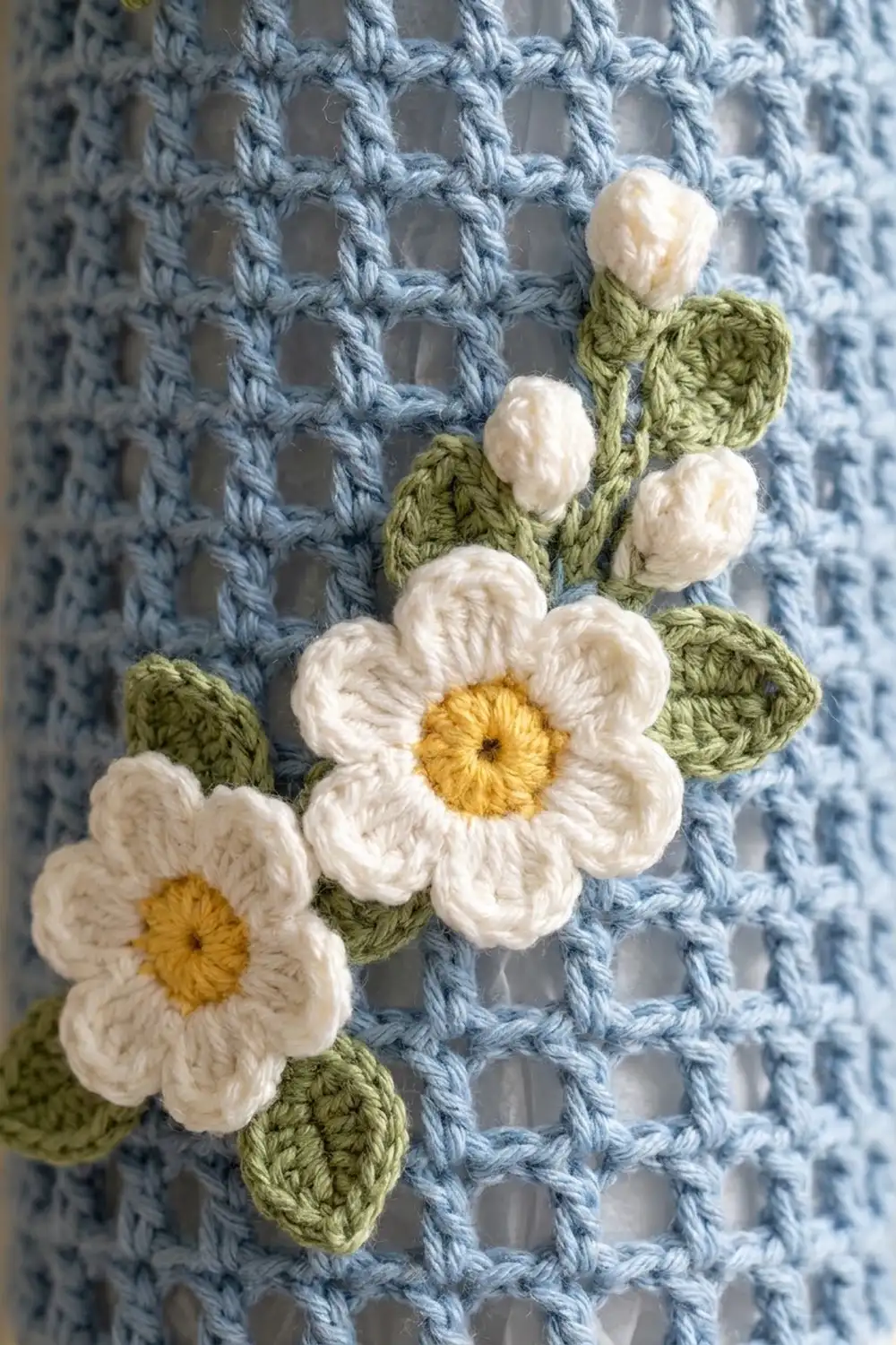 Crocheted white daisies with yellow centers and green stems arranged on a light blue knitted fabric background.