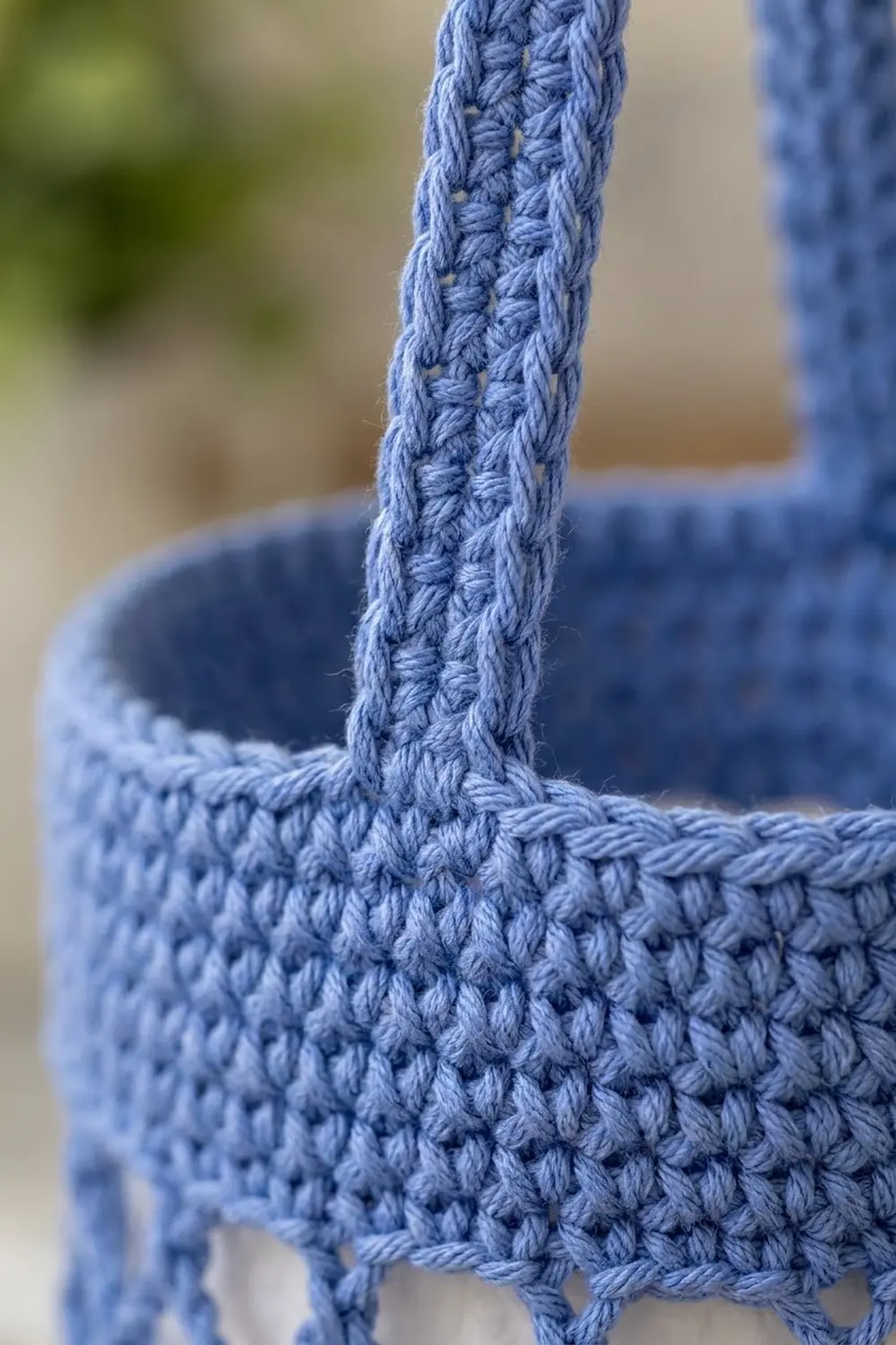 Close-up of a periwinkle blue crocheted or knitted tote bag with textured handles against a soft blurred background.