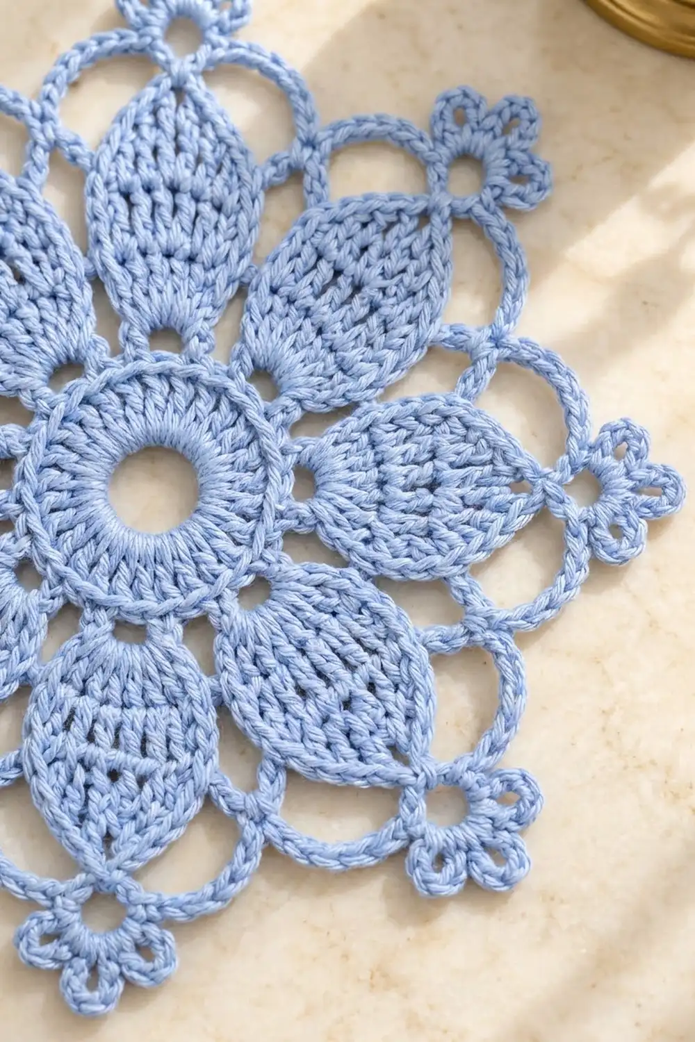 Light blue crocheted lace doily with intricate floral and leaf motifs displayed on a beige marble surface.