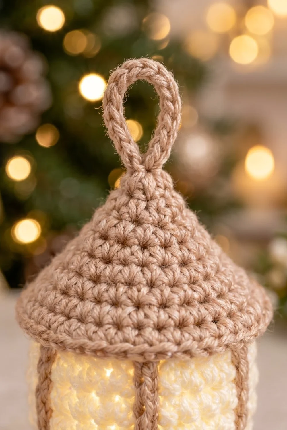 Crocheted beige bell ornament with rope loop hanging over warm glowing lights and blurred festive background.