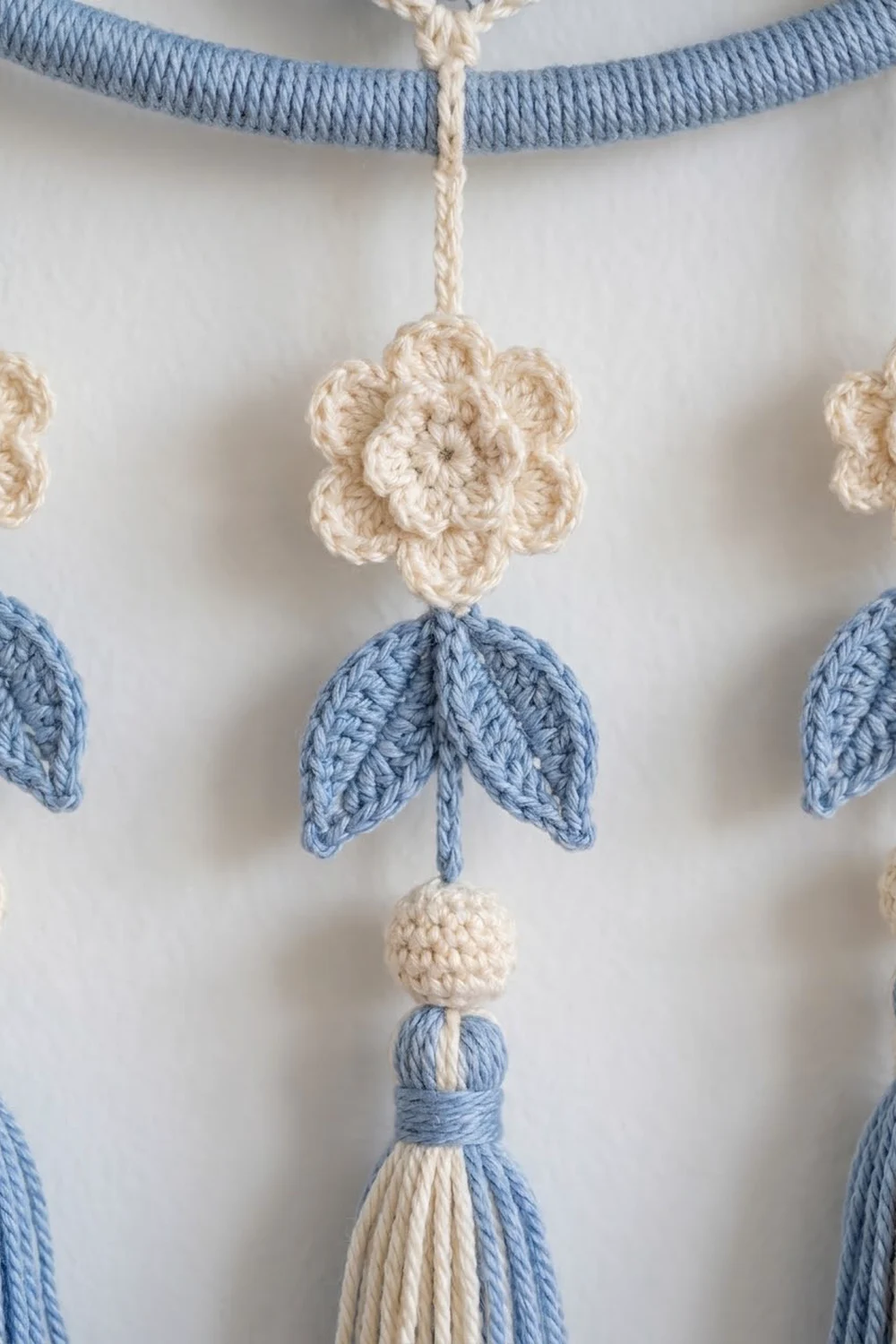 Close-up of crocheted flower mobile with cream blooms, blue leaves, and matching tassel details on white background.