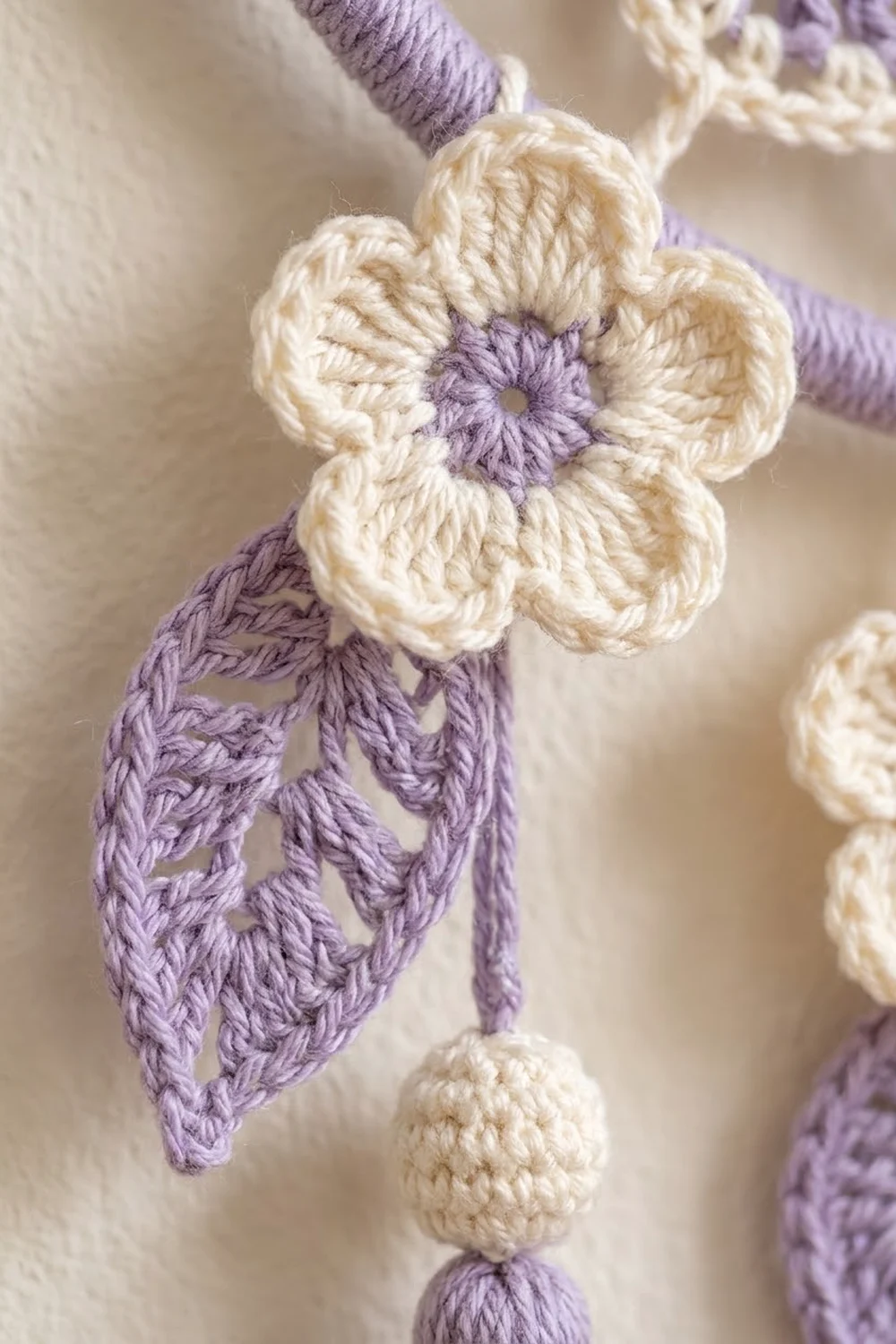 Crocheted flower ornament with cream petals, purple center, and matching leaf dangles in soft focus.