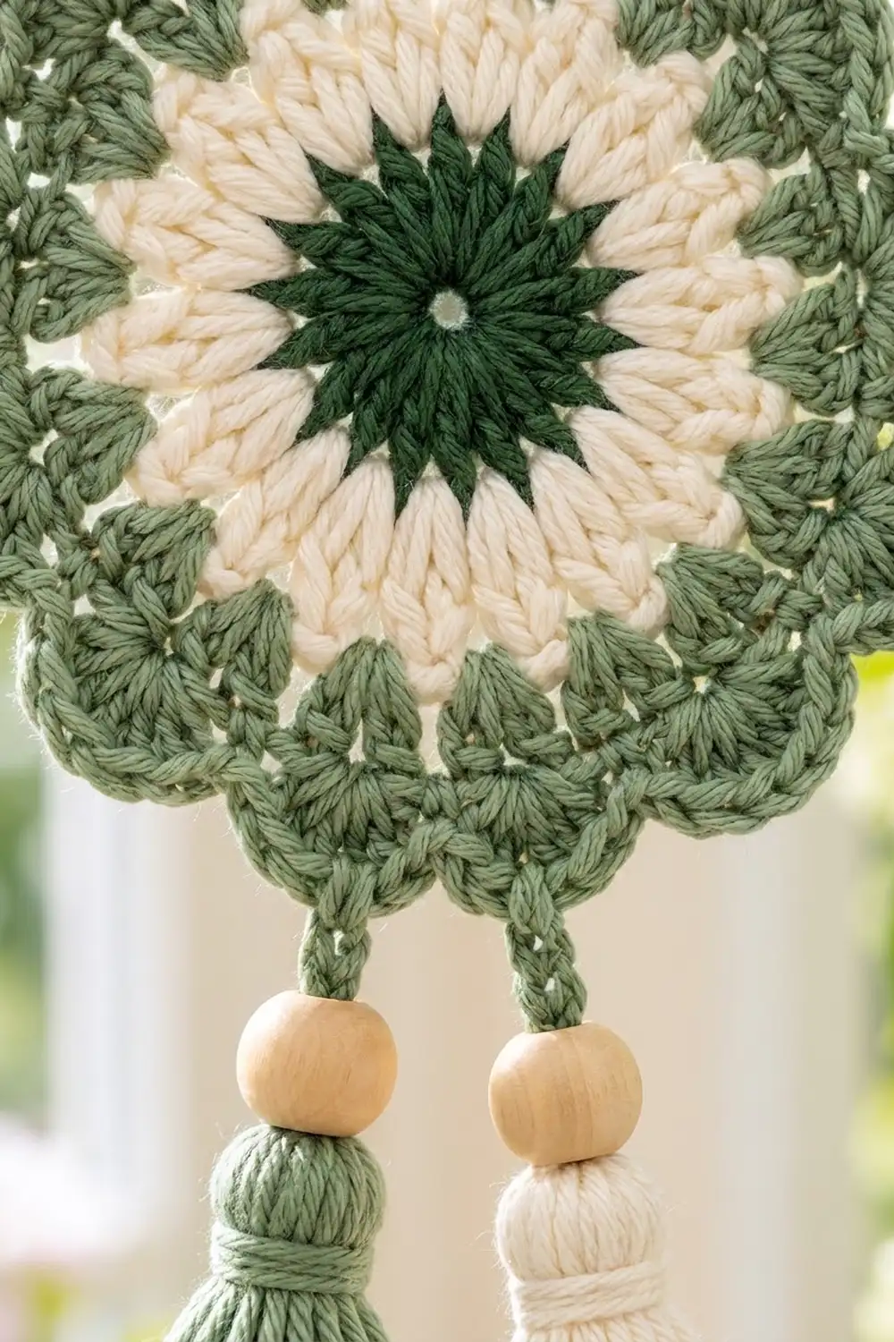 Crocheted flower mandala with sage green, cream, and dark forest green yarn featuring wooden bead tassel details.