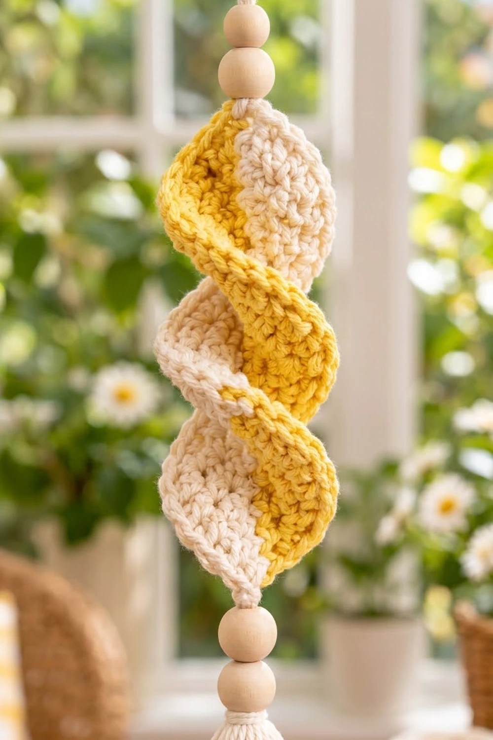 Crocheted beaded garland with alternating yellow and cream puffs hanging in sunny window with flowers behind.
