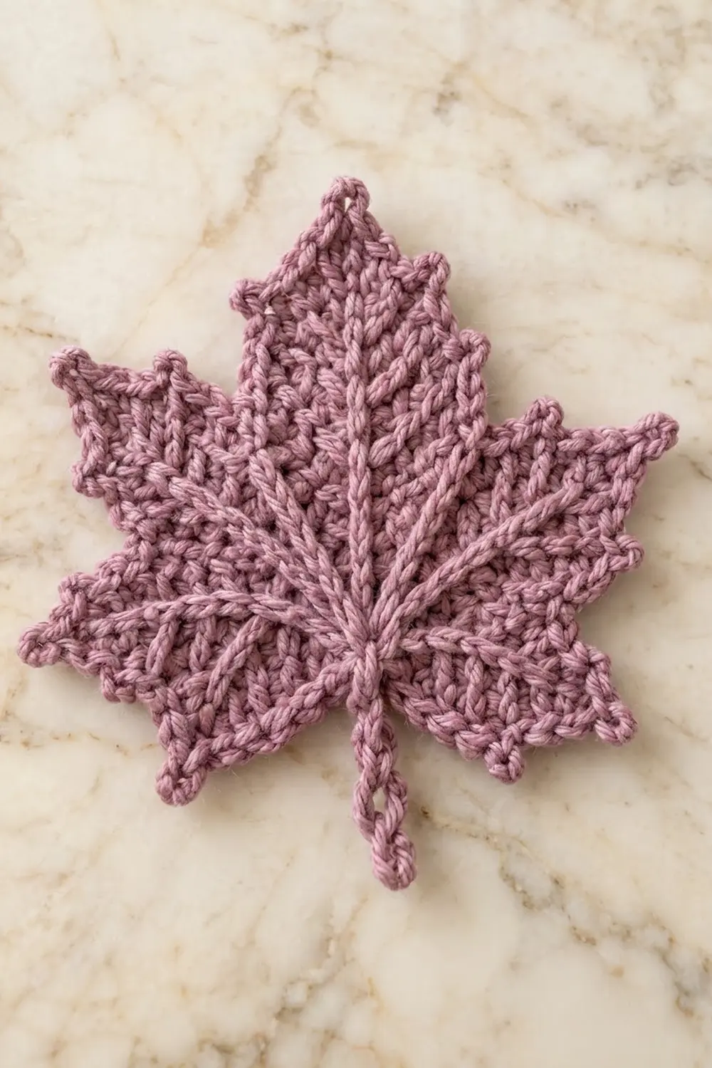 A dusty rose crocheted maple leaf with textured stitching rests on a cream marble surface.