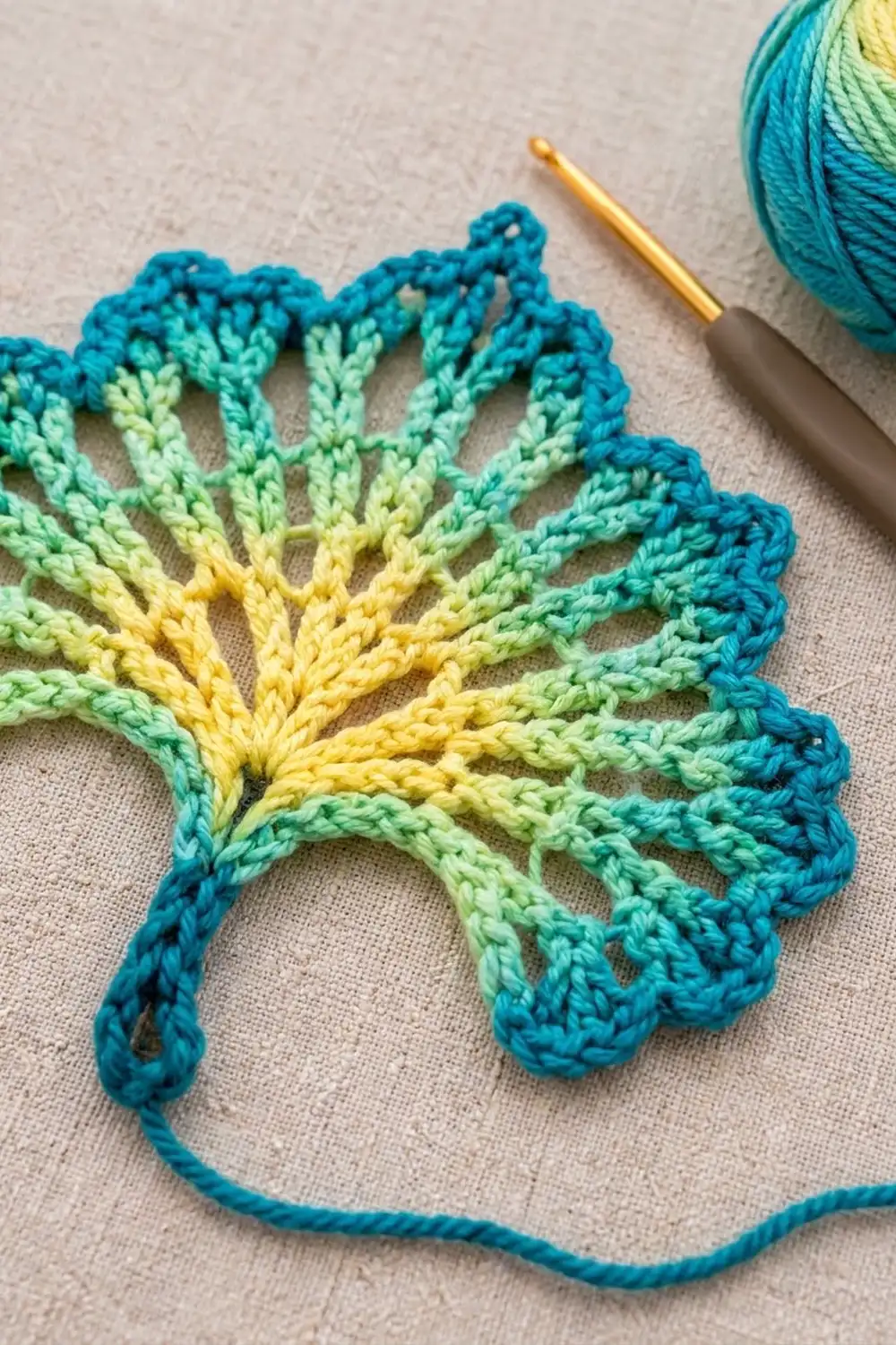 A crocheted leaf in gradient colors from teal to yellow lies on cream fabric beside a crochet hook and yarn.