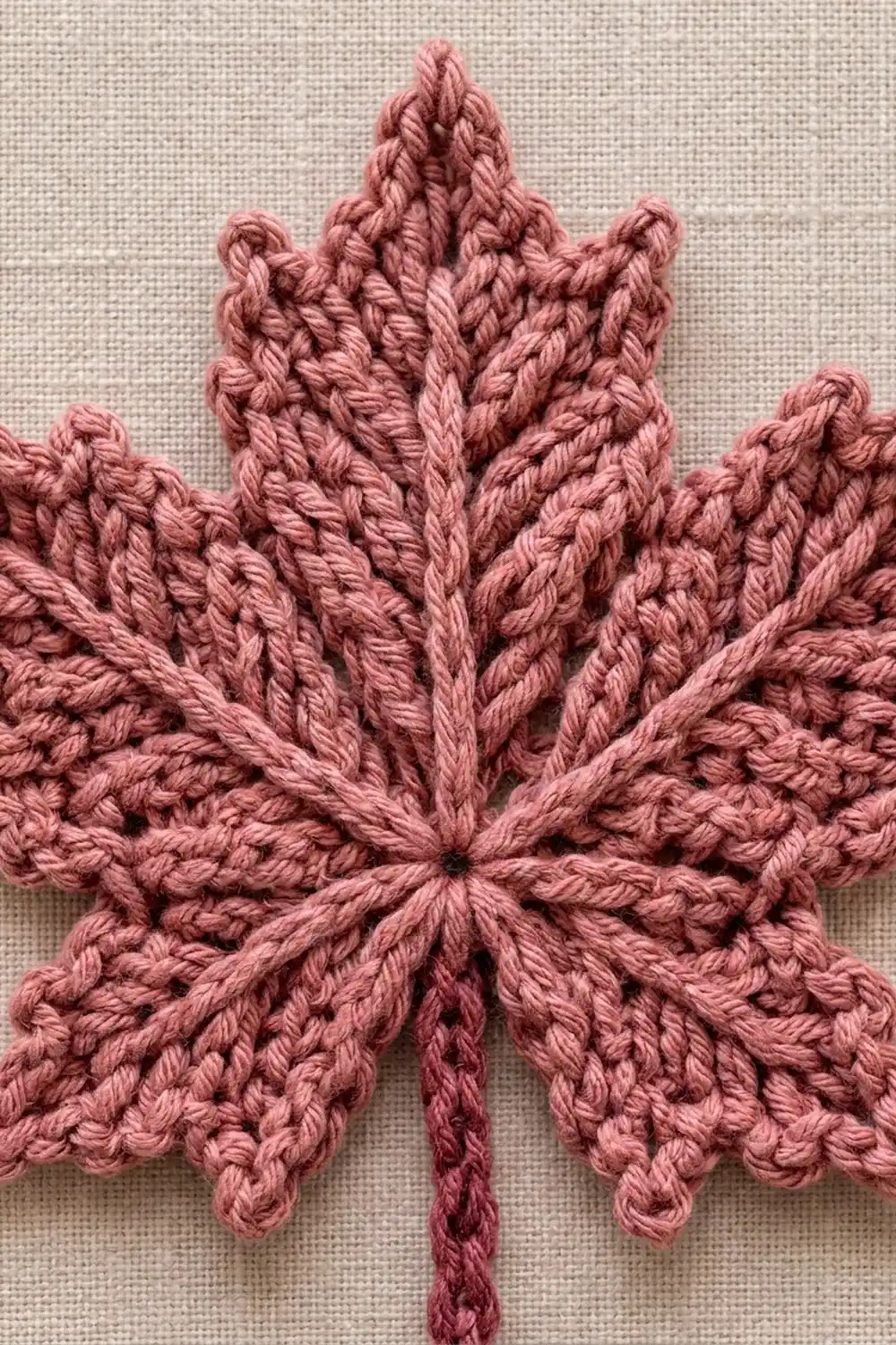 A dusty rose crocheted maple leaf appliqué displayed on a cream-colored linen fabric background.