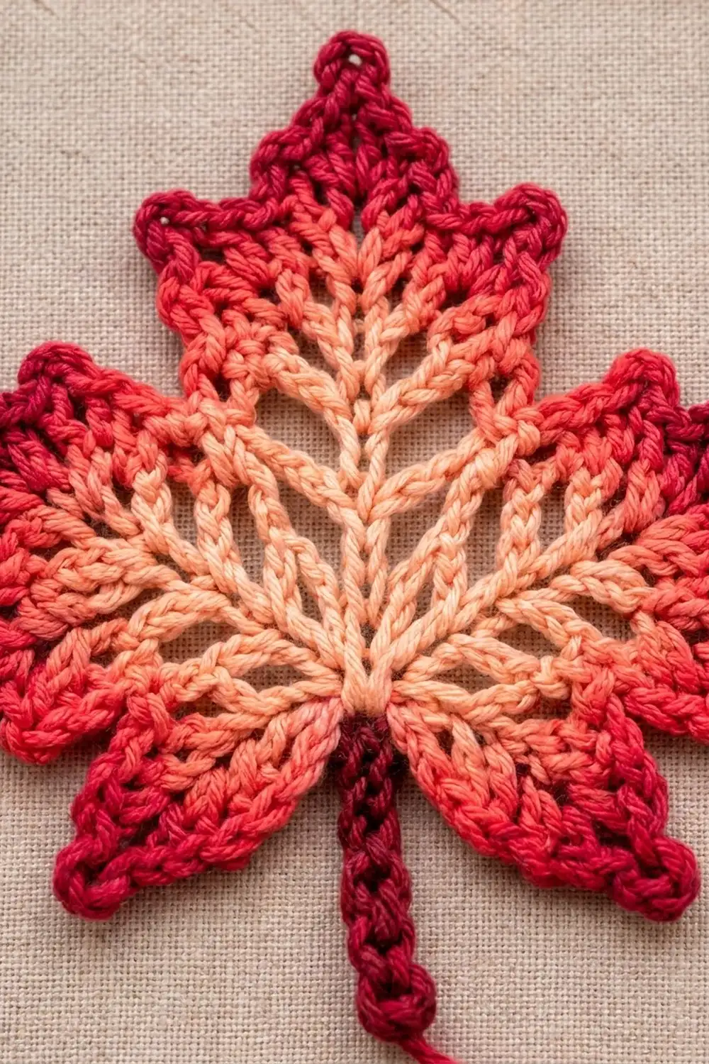 A crocheted maple leaf in red and coral gradient yarn displayed on beige linen fabric.