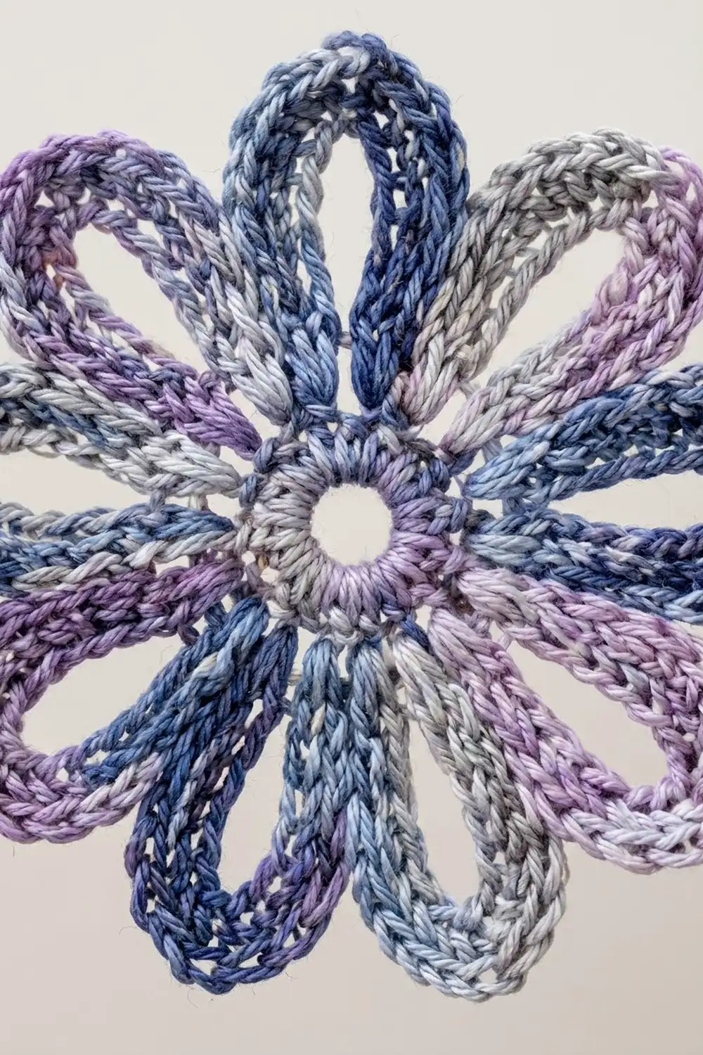 Braided yarn loops in shades of purple, blue, and gray arranged in a radial flower pattern against a cream background.