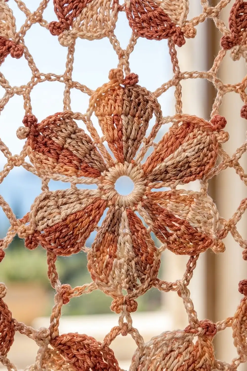 Close-up of handmade crochet lace with terracotta and cream flowers hanging against a window.