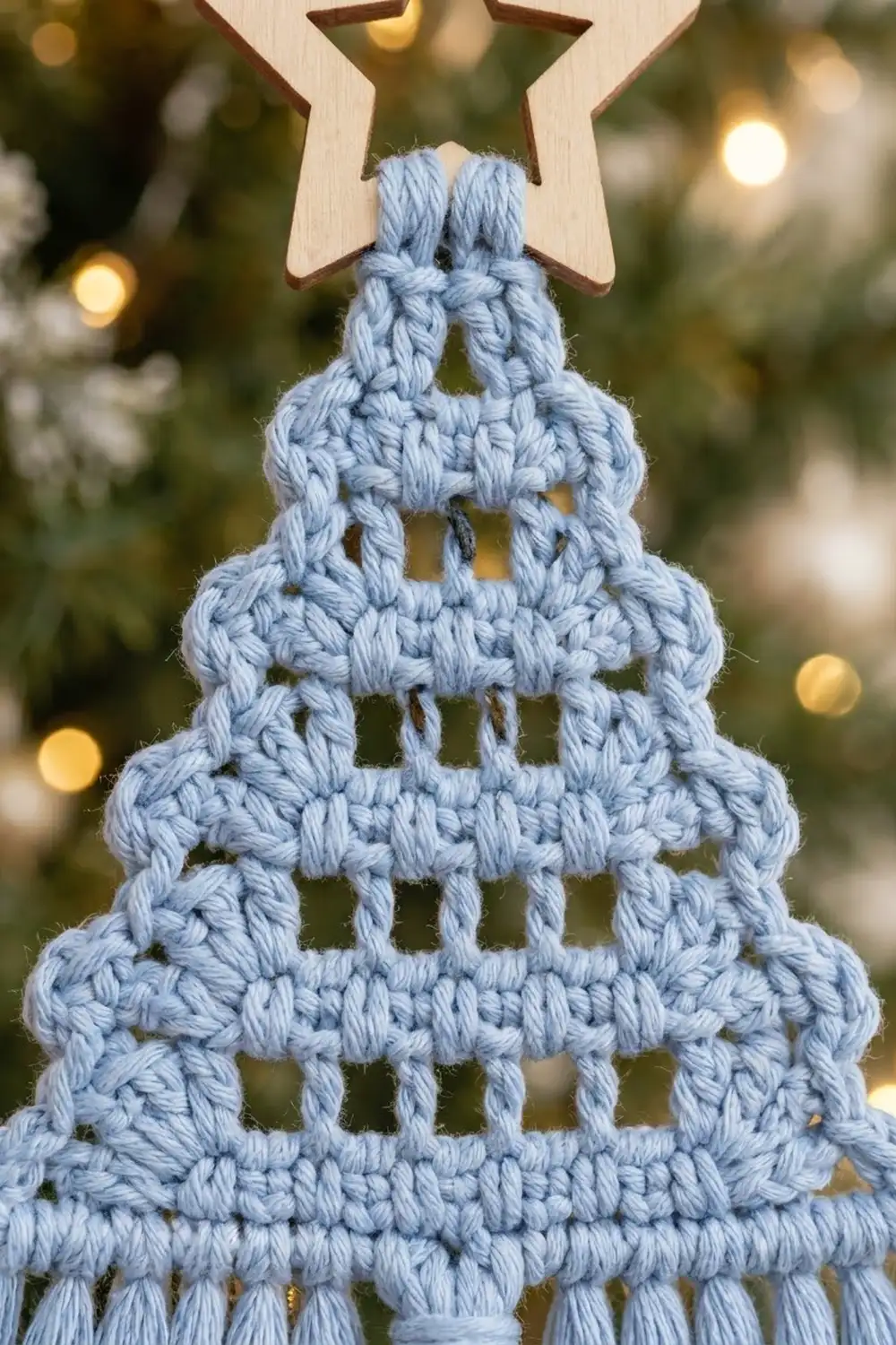 A light blue crocheted Christmas tree ornament with a wooden star topper hangs against a blurred festive background.
