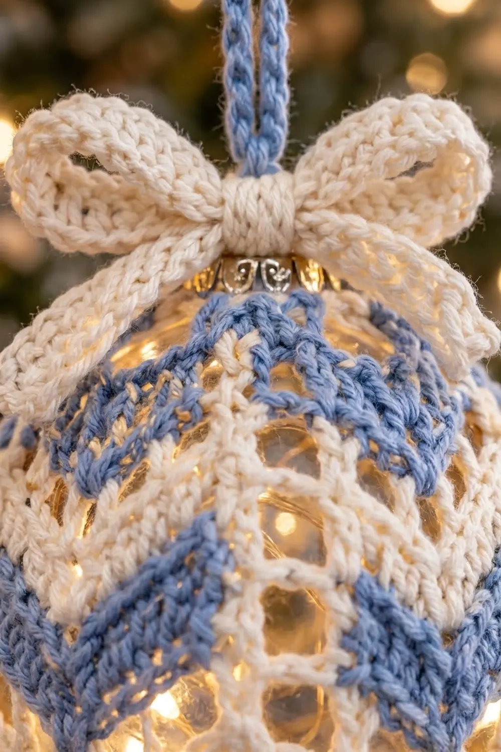 Crocheted ornament with cream and blue yarn featuring a bow top and gold accents against blurred lights.