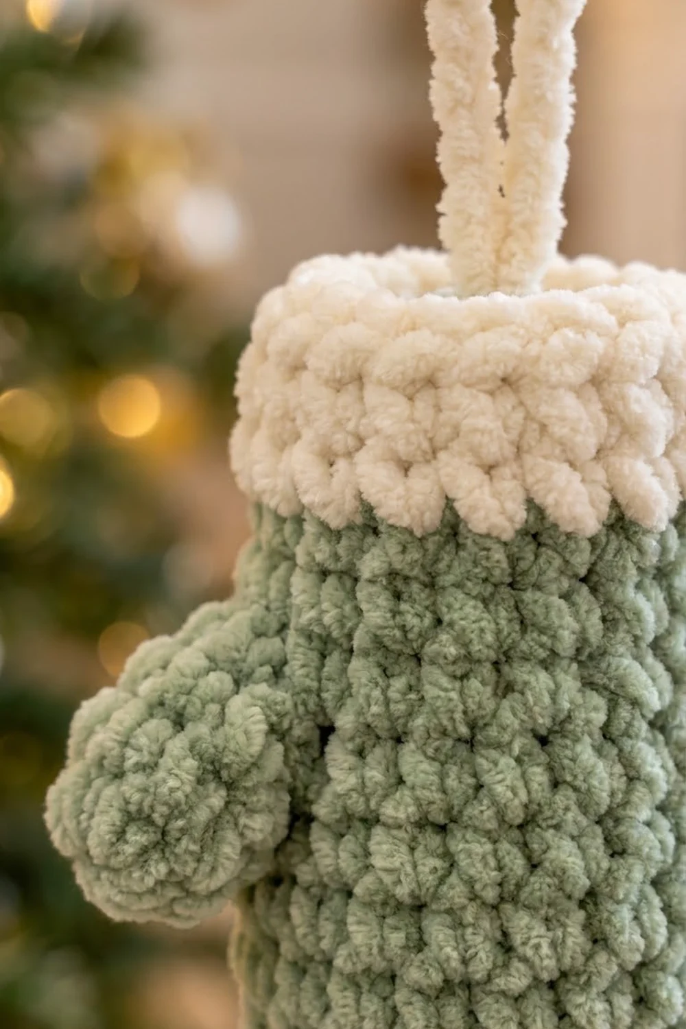A handmade crocheted Christmas stocking ornament in cream and sage green with textured bobble stitching and twisted cord loop.