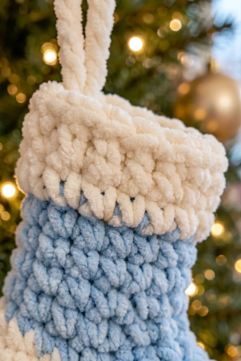 A handmade macramé stocking ornament in cream and blue hanging on a decorated Christmas tree with lights.