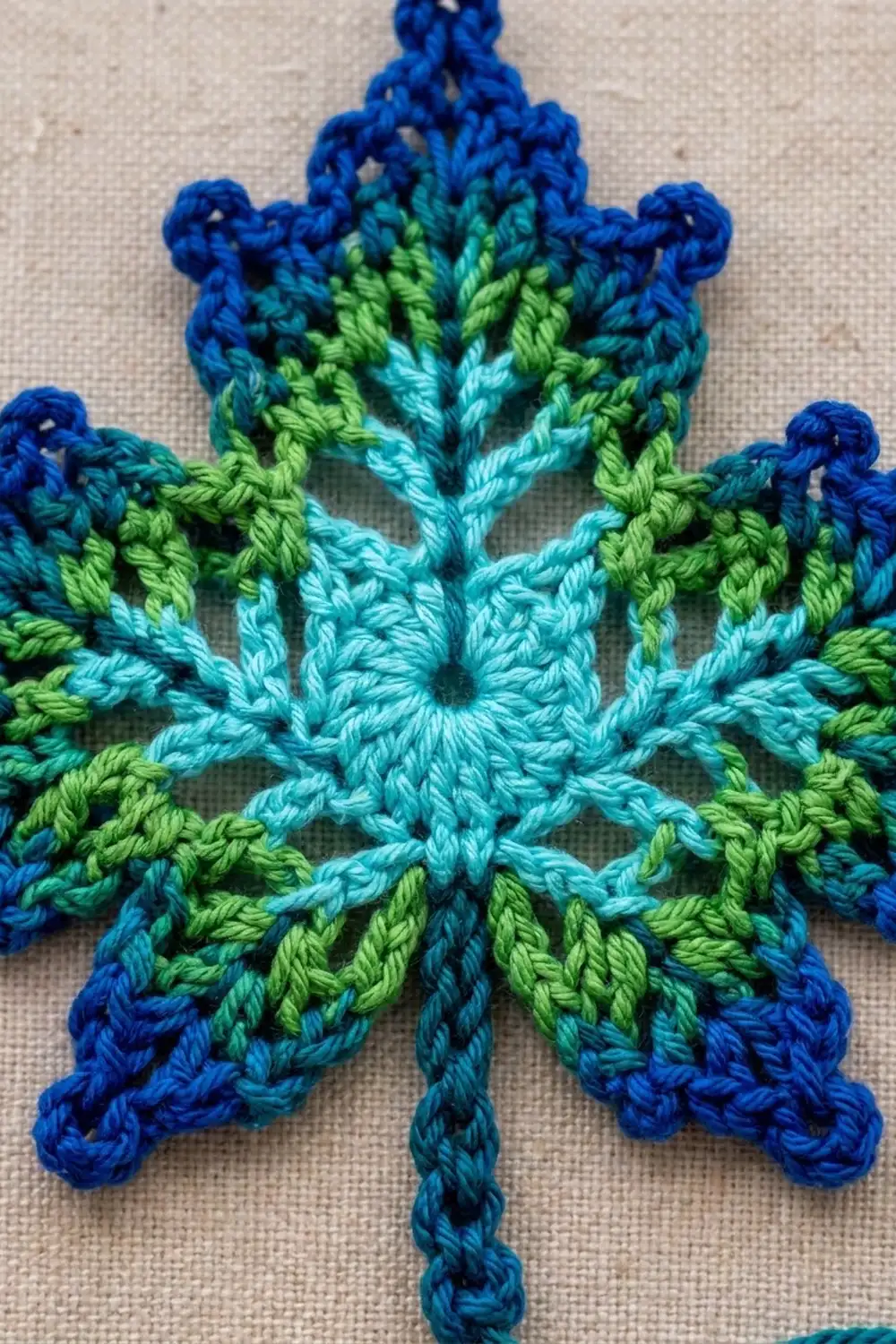 A crocheted snowflake ornament in blue, turquoise, and lime green yarn displayed on beige fabric.