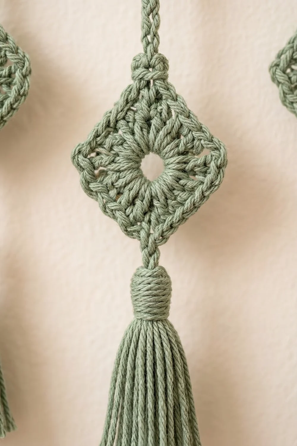 Close-up of a sage green crocheted diamond ornament with tassel hanging against a cream textured background.