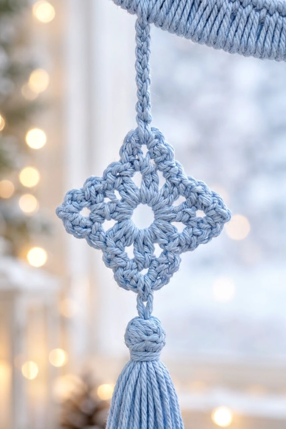 A light blue crocheted snowflake ornament with a tassel hangs against a snowy window with bokeh lights.