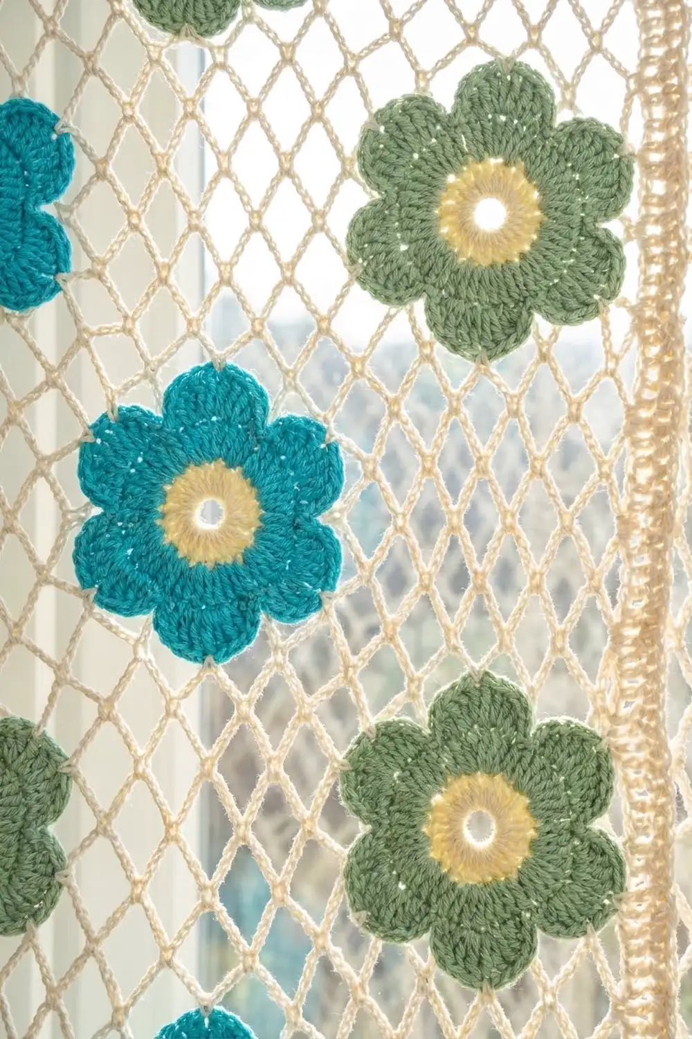 Crocheted flowers in teal and sage green with yellow centers attached to a beige macramé net curtain.