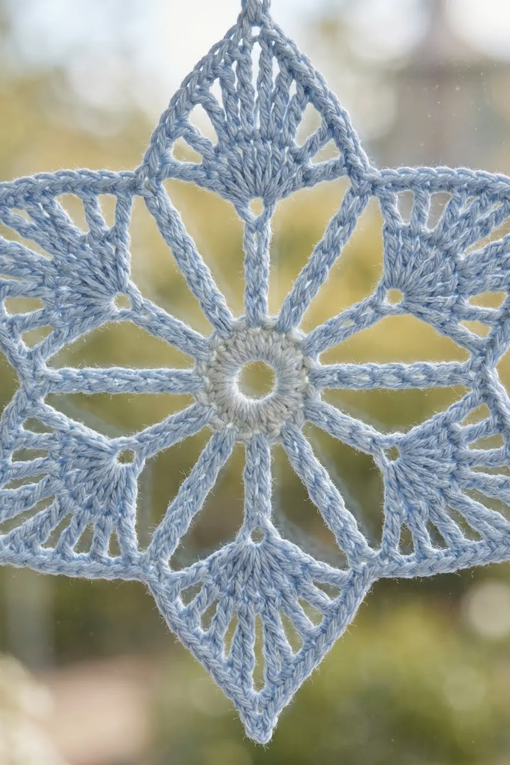 Delicate pale blue crocheted snowflake with white center displayed against blurred green and yellow outdoor bokeh.