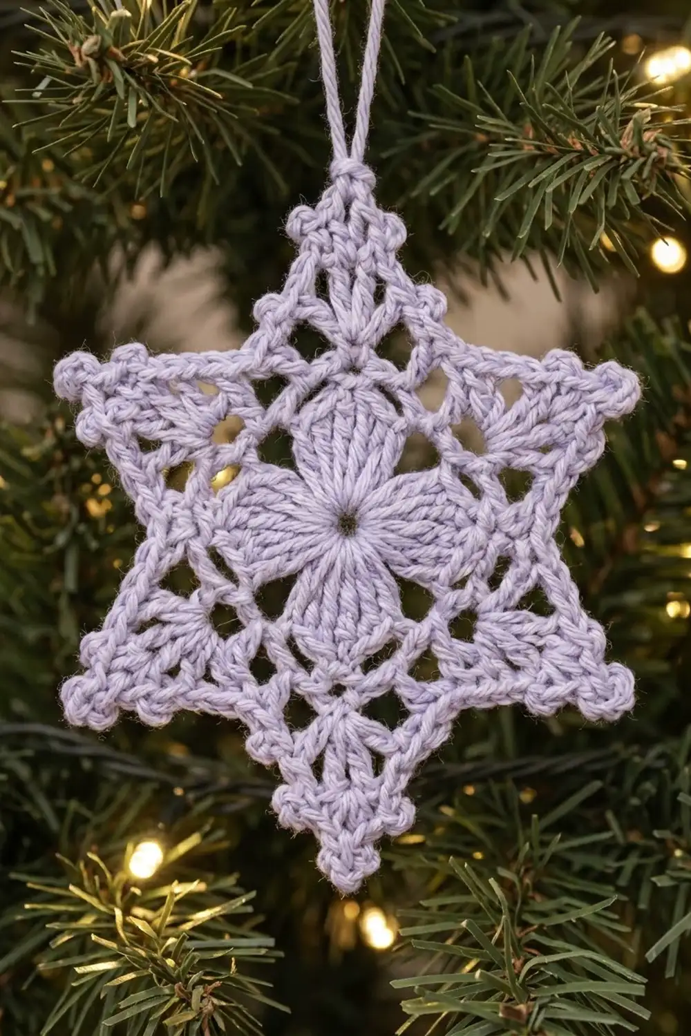 A lavender crocheted snowflake ornament with intricate lace patterns hangs from a pale ribbon on an evergreen tree branch.