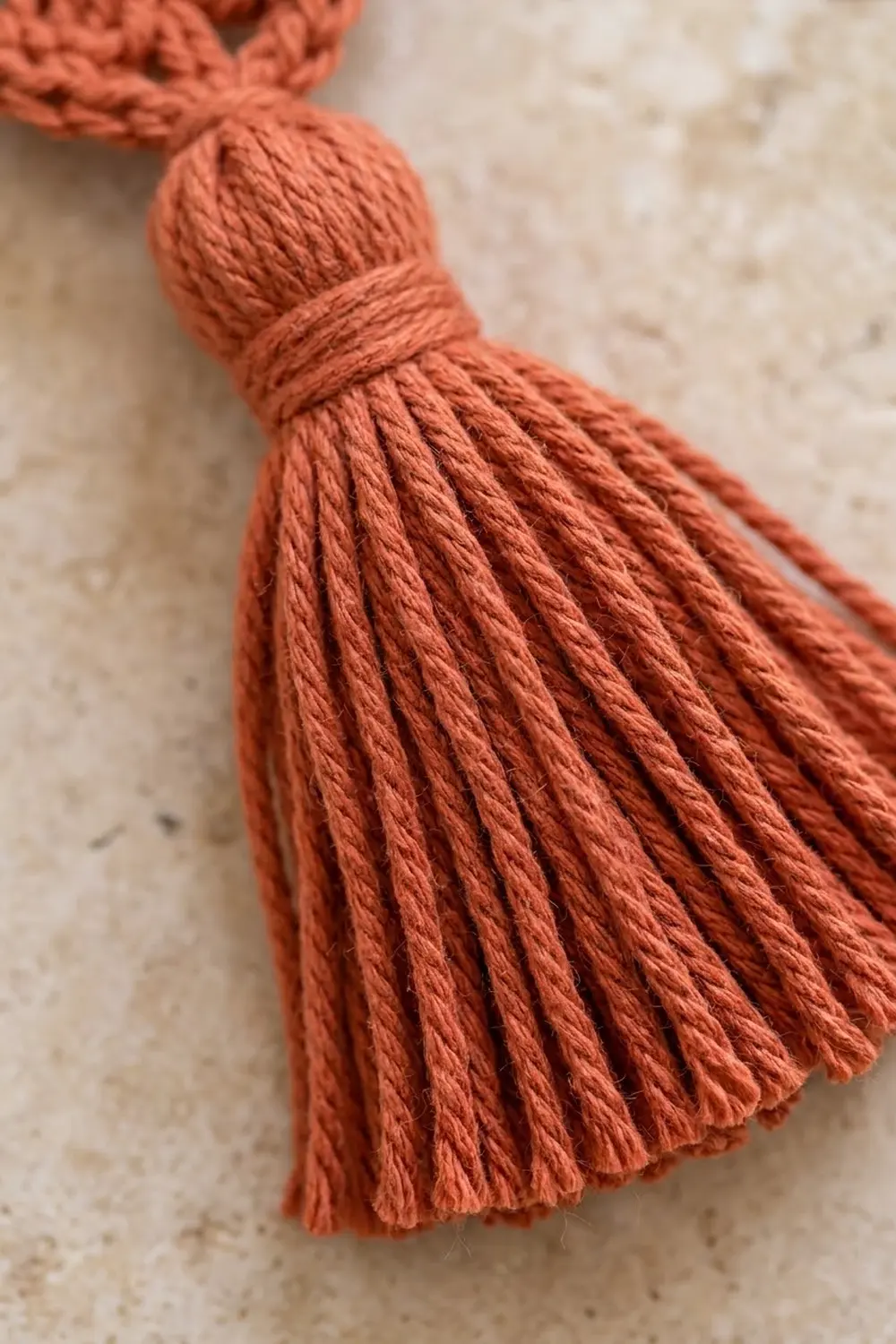 Rust-colored yarn tassel with twisted strands and bound knot displayed on neutral speckled linen.